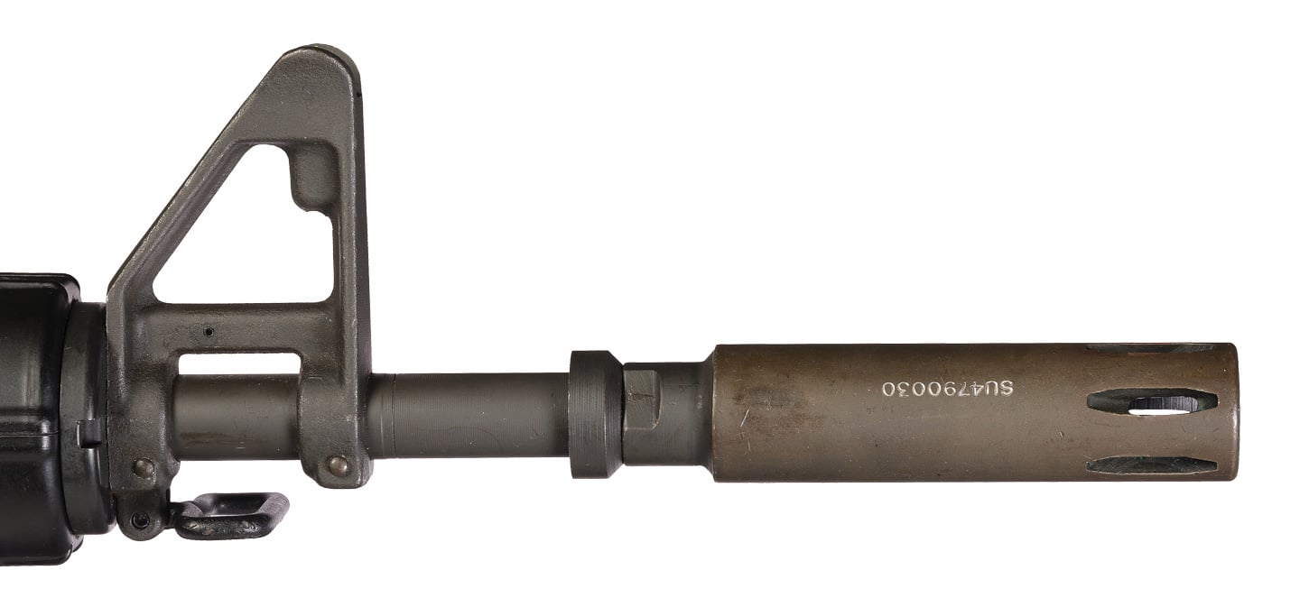 Close-up image of the XM177 moderator (muzzle device) mounted on a short-barreled CAR-15/XM177 carbine, photographed for Rock Island. The metal moderator is fixed to the muzzle and shows a smooth cylindrical exterior with a small exit port, designed to reduce visible muzzle flash and slightly lower the sound signature. The description emphasizes that the device is not user-disassemblable, making field cleaning difficult and causing performance to degrade as carbon and debris build up. The photo’s framing highlights the interface between the barrel and moderator and includes part of the short barrel and front sight for context. This alt text uses keywords such as XM177, moderator, muzzle device, CAR-15, flash suppression, and fouling to aid discoverability. It’s written to inform screen reader users about both the appearance and the functional limitations of the original XM177 moderator.