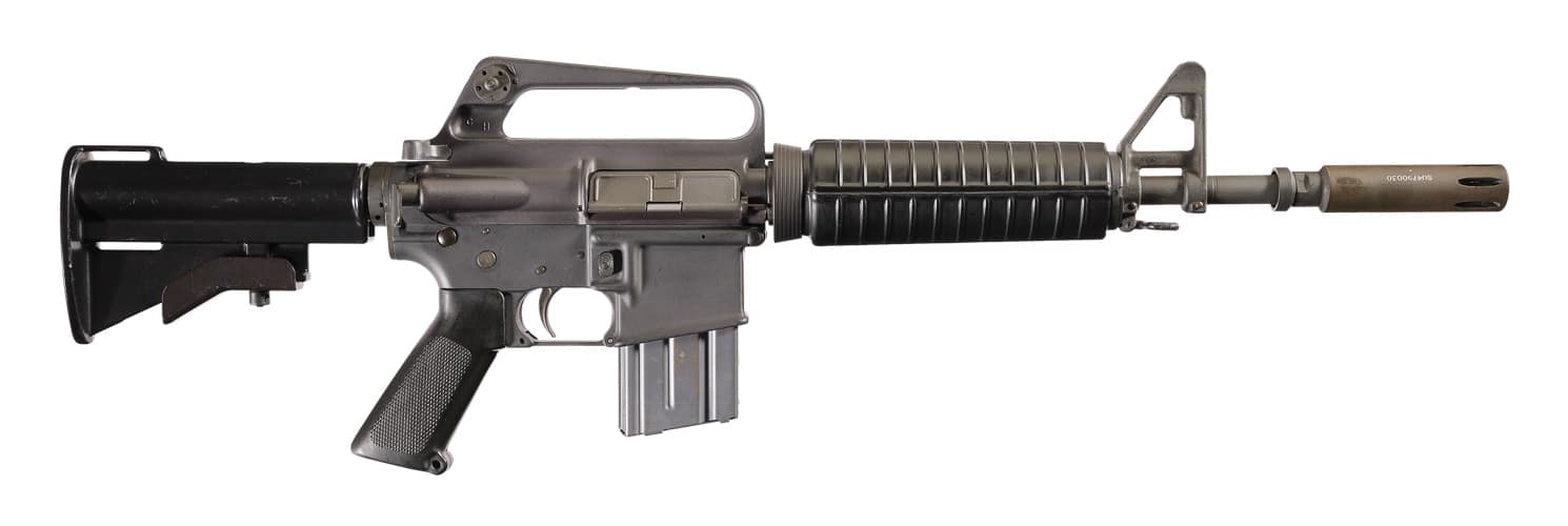 The image shows a typical XM177 rifle, also known as the Colt Commando or CAR-15 variant, as discussed in the article. The carbine features a short 10-inch barrel with a long moderator muzzle device, designed to reduce flash and change the weapon’s sound signature. A two-position collapsible aluminum stock extends from the receiver, reducing the rifle’s length from nearly 39 inches down to about 28 inches. A slim 20-round 5.56mm magazine is inserted, although later versions often used 30-round magazines. The overall design is compact, lightweight, and clearly intended for close-quarters combat and jungle mobility. XM177, Colt Commando, CAR-15, compact 5.56mm rifle, MACV-SOG weapon, and Vietnam War carbine for accessibility and SEO purposes.