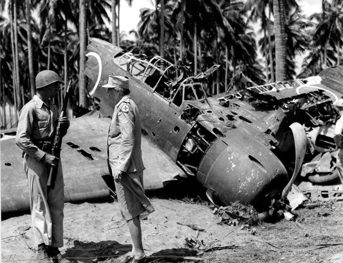 Black-and-white photograph of First Lady Eleanor Roosevelt examining the wreckage of a Japanese Mitsubishi A6M Zero fighter shot down on Guadalcanal. She is dressed in civilian attire, standing beside damaged aircraft parts as U.S. personnel look on. The image captures her historic wartime visit to the South Pacific, where she met with troops and inspected battlefields. The destroyed Zero symbolizes the fierce aerial combat that took place during the Guadalcanal campaign. Roosevelt’s presence on the island was both symbolic and morale-boosting, demonstrating the home front’s connection to the front lines. The photo highlights her unique role as an active First Lady during World War II.