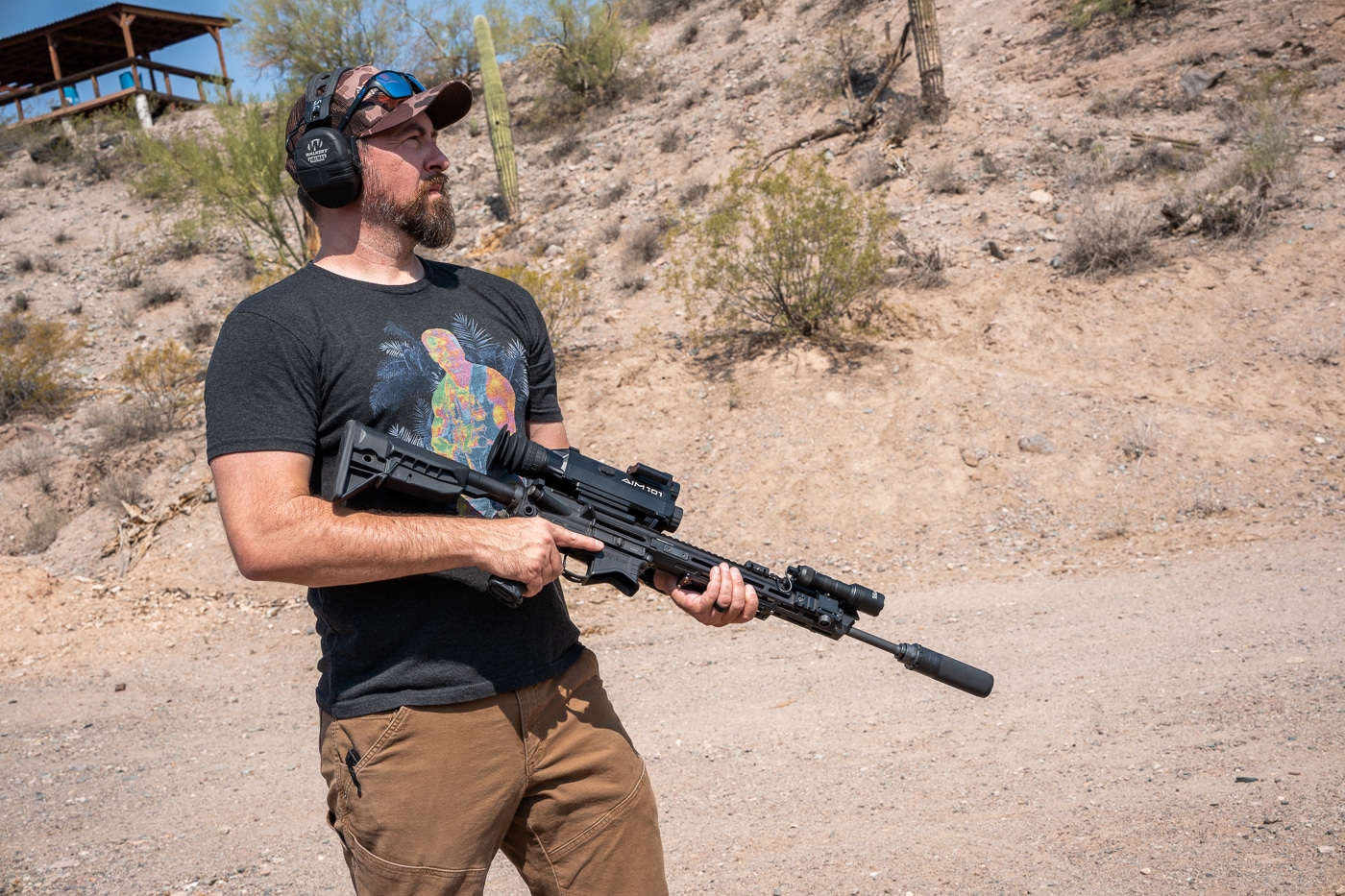 The photo shows the author carrying a Springfield Armory SAINT rifle equipped with the Shinenyx AIM-101 thermal and night vision optic. The rifle is held at the ready position while the author trains on an outdoor shooting range. The AIM-101 is visible on the top rail, with its compact housing and rubberized controls standing out. The optic’s size integrates well with the AR-15 platform, allowing smooth movement during drills. Range features, including berms and target stands, are visible in the background. The setup illustrates how the AIM-101 remains stable and balanced while in motion. This demonstrates the optic’s suitability for real-world shooting scenarios where both mobility and durability matter.