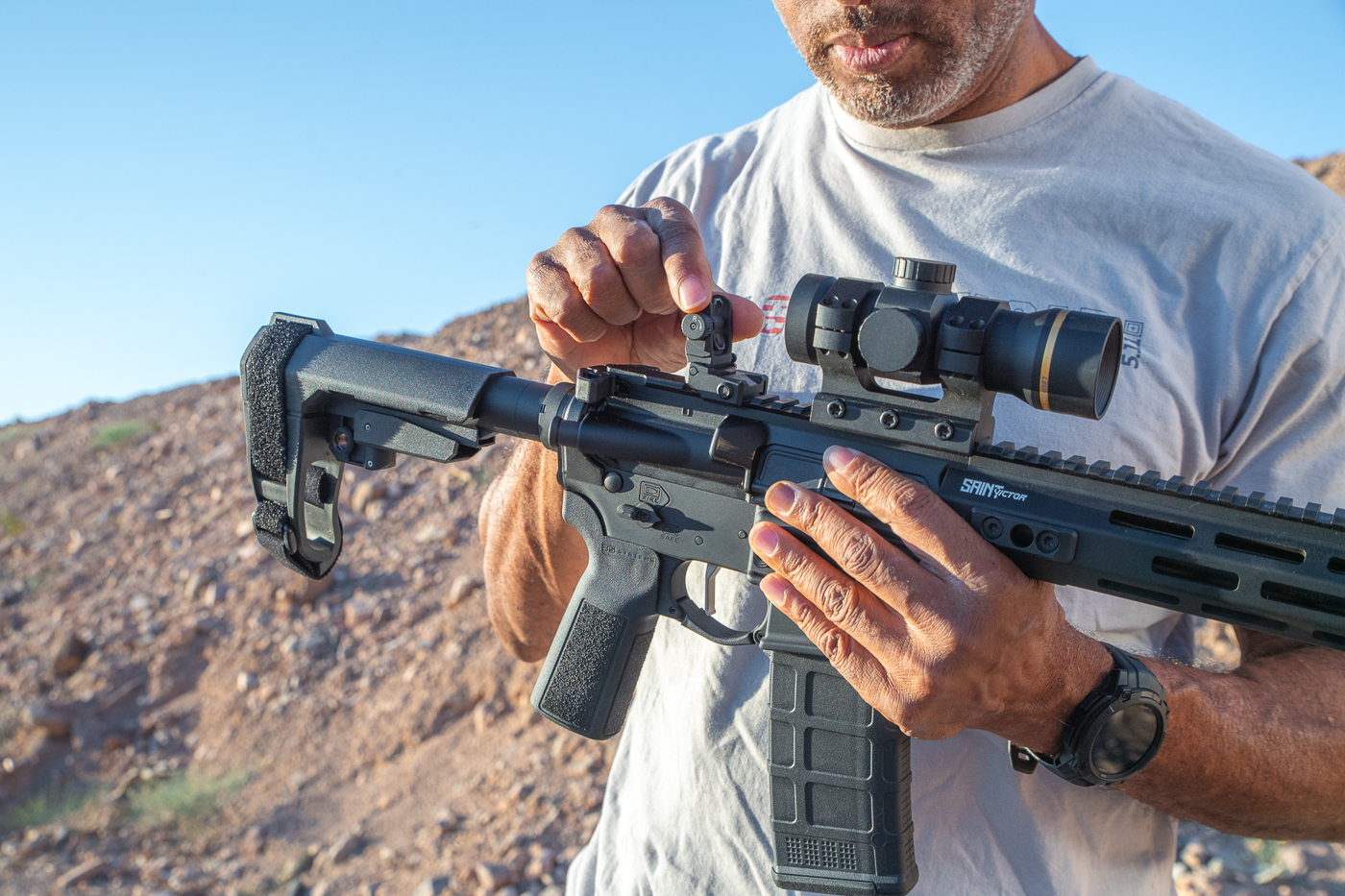 The image shows a shooter demonstrating how to deploy the iron sights on the Springfield Armory SAINT Victor 2 rifle. The shooter is manually flipping up the low-profile sights, which are mounted on the rifle’s full-length Picatinny top rail. These new sights are designed to sit lower than the original SAINT Victor units, providing a sleeker fit and less interference with optics. The photo captures the shooter’s hands manipulating the sights, illustrating how quickly they can be engaged. The rifle’s other features, such as the free-float handguard with M-Lok slots and the B5 Systems furniture, are also visible. The iron sights are made of aluminum for durability and include windage adjustments on the rear unit. This image emphasizes the practical role of backup sights for shooters who want redundancy alongside red dots or scopes.
