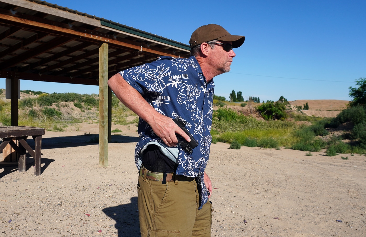 Action photo of the reviewer at an outdoor shooting range drawing a Springfield Hellcat Pro pistol from the StealthGearUSA Ventcore 2.0 IWB Mini holster. The holster is positioned at the 4 o’clock carry spot on the hip, worn inside the waistband. The author is mid-draw, with the pistol partially out of the molded Kydex shell. The padded Ventcore backing is pressed against the body, showing how the holster stays in place during movement. The scene demonstrates retention, comfort, and draw speed in a realistic training scenario. Dust, grass, and range targets are visible in the background.
