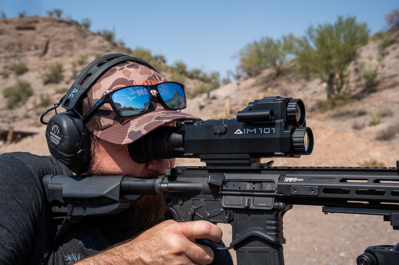 The photo shows the author firing a Springfield Armory SAINT rifle with the Shinenyx AIM-101 mounted on top. The rifle is shouldered, and the shooter is engaged in live-fire testing. The optic’s eyepiece is aligned with the shooter’s eye, showing how it is used in real-time. The sturdy housing and rubberized buttons of the AIM-101 are visible along the top. The scene takes place outdoors on a flat range under bright daylight conditions. The optic’s display is designed to remain visible even in direct sunlight, which was part of this test. This setup demonstrates how the AIM-101 performs under recoil and maintains zero while providing thermal and night vision fusion modes.