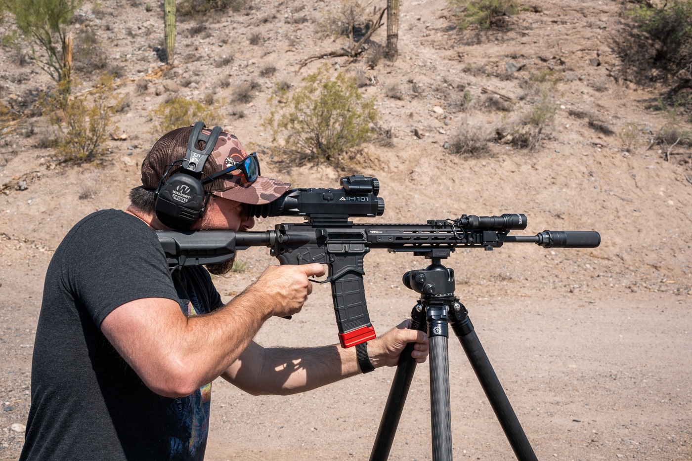 The photo shows the author at a shooting range using a Springfield Armory SAINT rifle equipped with the Shinenyx AIM-101 optic. The rifle is secured on a tripod, providing stability during testing. The author is positioned behind the rifle, sighting through the AIM-101’s eyepiece. The scope’s housing, top-mounted controls, and rear display are visible in use. The tripod setup reduces movement, allowing for precise adjustments and accurate evaluation of the optic. The background includes range lanes and targets, emphasizing the live-fire environment. This scene demonstrates how the AIM-101 performs during controlled testing with thermal, night vision, and fusion imaging capabilities.
