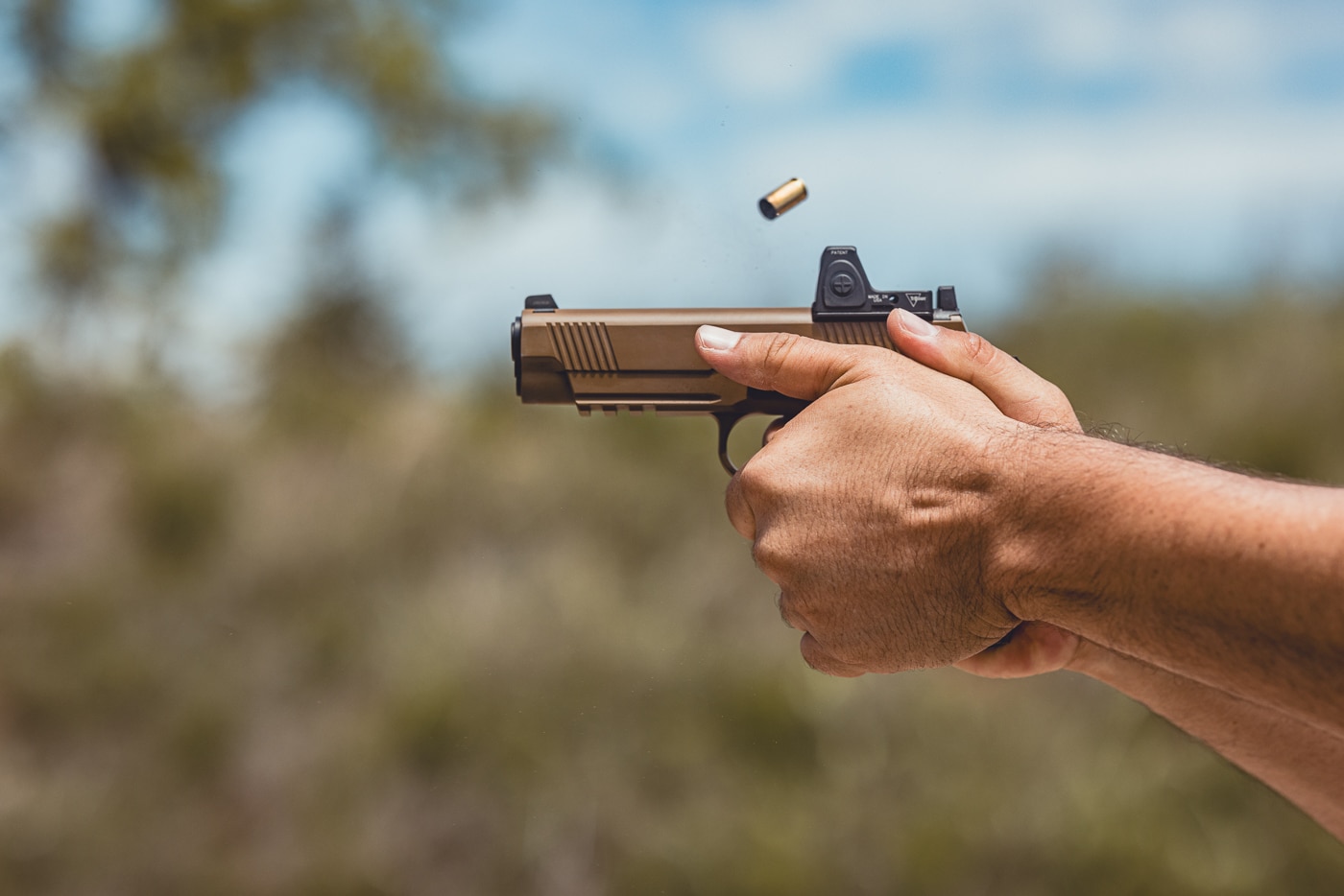 This close-up image shows the author shooting a Springfield Armory 1911 TRP AOS equipped with a red dot optic mounted directly on the slide. The pistol is in firing position, and the optic’s compact design integrates seamlessly with the 1911’s profile. The shooter maintains a firm two-handed grip, demonstrating recoil control and proper stance. Visible details include the forged slide, front serrations, and textured grips of the TRP series. The mounted red dot emphasizes the Agency Optic System’s ability to bring modern technology to a century-old design. This photo illustrates how the TRP AOS performs under live fire with an optic attached.