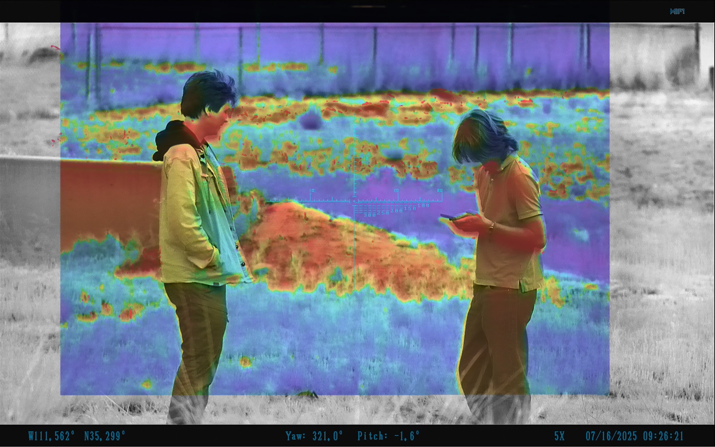 The image is a captured view through the Shinenyx AIM-101 fusion optic showing two people. Their bodies are outlined with thermal highlights, making them appear bright compared to the cooler background. Digital night vision detail provides context, including the ground and surrounding environment. The contrast between the thermal overlay and night vision feed makes it easy to identify the individuals. Both figures stand out clearly, showing movement and body positioning. This demonstrates how the AIM-101 allows for quick detection and confirmation of human presence. The FusionView example emphasizes the scope’s usefulness for hunting, tactical operations, and search and rescue.