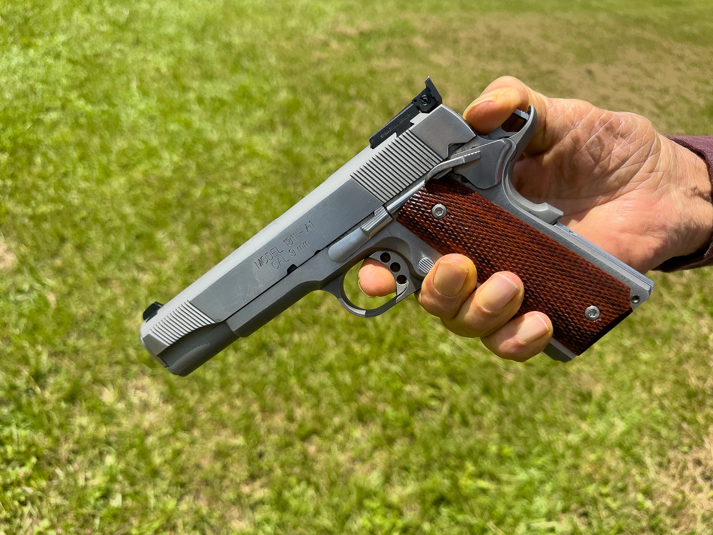 The image shows Massad Ayoob holding a 1911 pistol while demonstrating how to lower the hammer on an empty chamber. His thumb is placed on the hammer, illustrating how to control its movement while pressing the trigger. The firearm is angled in a way that makes the hammer, slide, and grip safety clearly visible. Ayoob emphasizes that this action should only be done after confirming the gun is unloaded multiple times. His expression and stance indicate an instructional setting rather than active shooting. The photo highlights safe gun handling for storage or administrative handling of the 1911. It captures the fine motor control required to safely lower the hammer without risk of discharge.