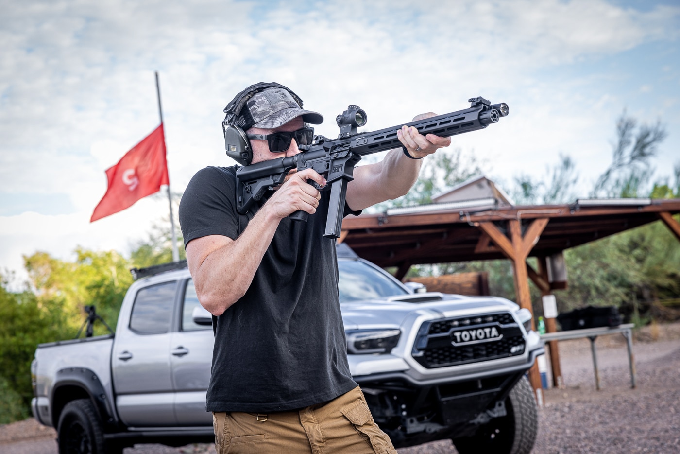 Male shooter firing a carbine rifle at an outdoor shooting range, demonstrating the compact firearm's maneuverability and ease of handling compared to full-length rifles. The carbine's shortened barrel length and reduced overall dimensions make it ideal for tactical shooting and sport shooting applications. Shooter maintains proper stance while operating the lightweight carbine, showcasing how these firearms offer improved control and faster target transitions. The image illustrates typical carbine use in civilian shooting sports and training scenarios. Modern carbines like this serve multiple purposes including home defense, competitive shooting, and recreational range use. The firearm's compact profile allows shooters to quickly move between firing positions while maintaining accuracy at close to medium ranges. This type of shortened rifle design has become increasingly popular among sport shooters and firearms enthusiasts for its versatility and practical handling characteristics in various shooting environments.