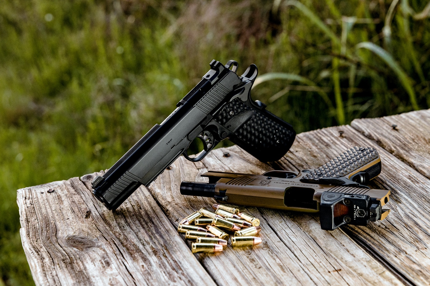 This image shows two Springfield Armory TRP 1911 AOS pistols placed on a shooting bench. The pistol on the left is equipped with a modern red dot sight, while the pistol on the right retains traditional iron sights. Both pistols are full-size 1911 handguns, built with forged frames and featuring Springfield’s signature aggressive checkering and grips. The setup demonstrates the flexibility of the Agency Optic System, which allows shooters to easily swap between optics and irons. The photo emphasizes the contrast between a classic 1911 setup and one upgraded with modern optics. It reflects the balance of tradition and innovation that defines the TRP AOS.