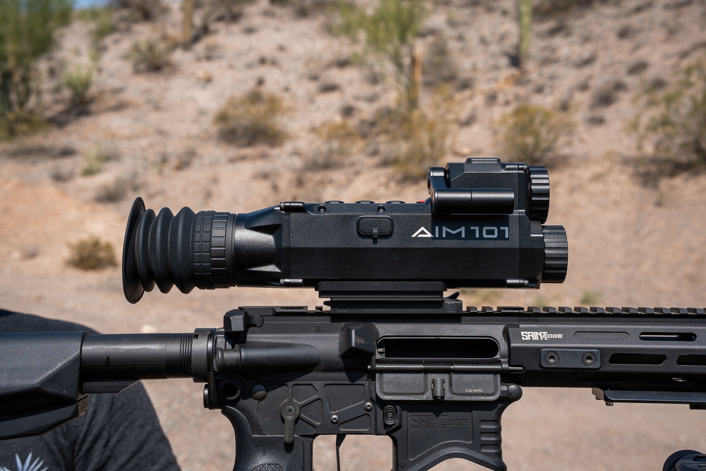 The image shows a side profile of the Shinenyx AIM-101 fusion optic. The rugged all-metal housing is visible, with clean lines and a matte finish. A scroll wheel is located on the side, used for menu navigation and zoom adjustment. The mounting base sits along the bottom of the optic, designed for attachment to a Picatinny rail. The eyepiece extends from the rear, positioned for comfortable alignment with the shooter’s eye. The design appears compact and well-balanced for both handheld and weapon-mounted use. This angle emphasizes the AIM-101’s durability, compact footprint, and field-ready build.