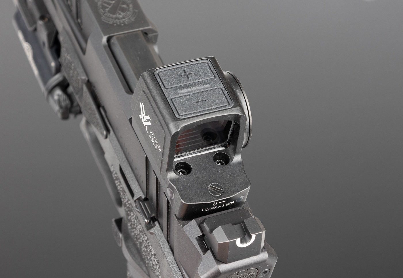 Close-up photo of the top housing of the Vortex Venom Enclosed red dot sight showing the two brightness adjustment buttons. The buttons are recessed into the optic’s aluminum housing, labeled for increasing and decreasing brightness. These controls allow the user to cycle through 12 total brightness levels, including two that are compatible with night vision devices. The positioning on top of the sight ensures they are easy to reach without blocking the view through the optic. The image emphasizes the design’s balance of accessibility and durability for both defensive and range applications. This detail shot helps readers understand the practical controls built into the Venom Enclosed, as described in the review.