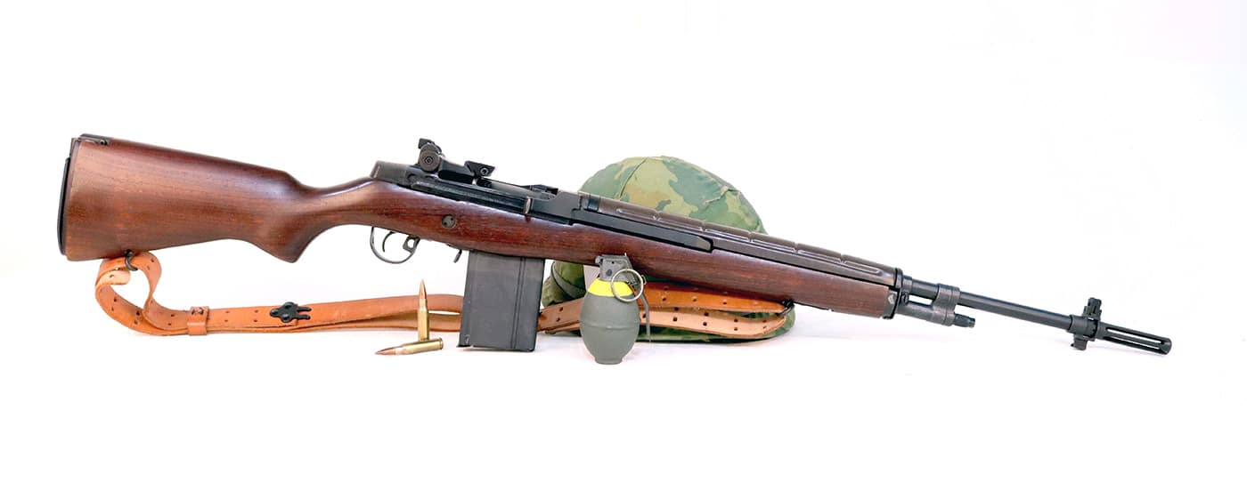 A detailed photo of the Springfield Armory M1A rifle, shown in profile to highlight its resemblance to the original M14. The gun features a walnut stock, parkerized metal finish, and a distinctive operating rod handle on the right side. Its layout and lines clearly reflect the M14’s military heritage. The M1A maintains the same gas-operated action and traditional trigger group, though configured for civilian use. The lighting emphasizes the craftsmanship and texture of the stock, showing its classic service-rifle form. This image visually connects the M1A to its M14 origins and helps illustrate how Springfield Armory adapted a combat-proven design for the modern marksman. It reinforces the rifle’s reputation for durability, accuracy, and historical significance.