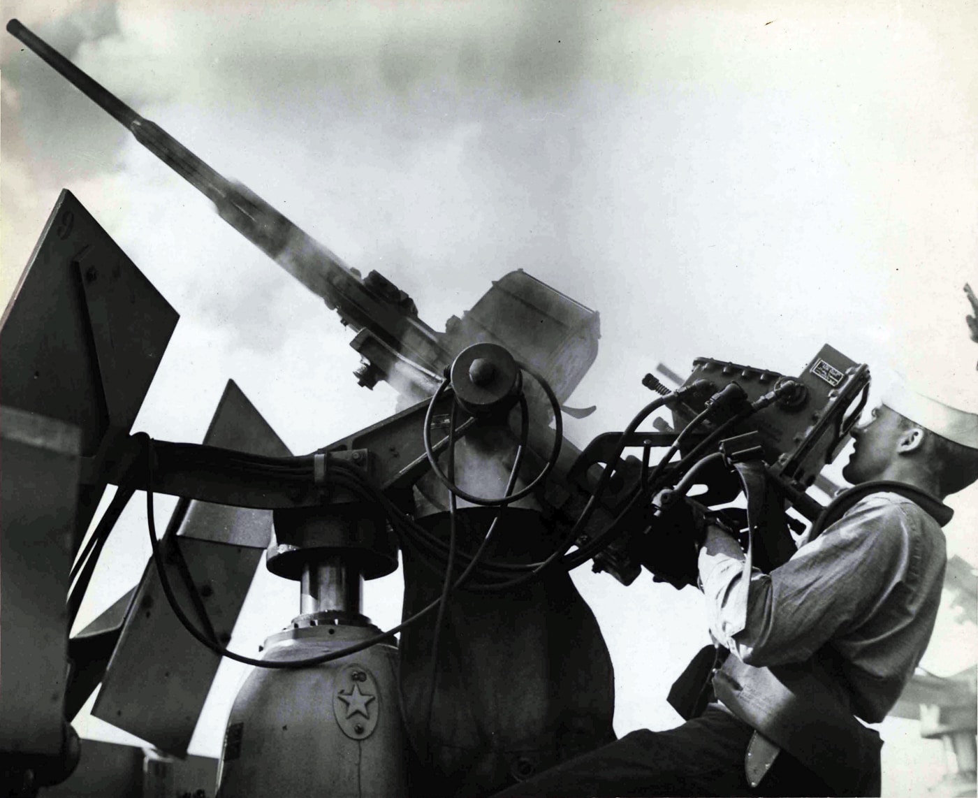 A black-and-white photo showing a 20mm Oerlikon anti-aircraft gun crew aboard the USS Natoma Bay (CVE-62) during World War II. The gunner stands behind the shoulder rests, aiming the weapon skyward, while a loader holds a 60-round drum magazine ready to feed the gun. The curved gun shield partially surrounds the mount, offering protection from shrapnel and wind. The flight deck and ocean horizon are visible behind the men, suggesting active operations in the Pacific. The photo conveys the tension of combat readiness as Japanese kamikaze aircraft threatened the escort carrier. The Oerlikon’s compact design and fast firing rate made it one of the Navy’s most widely used anti-aircraft guns during Japanese Kamikazes in World War II.