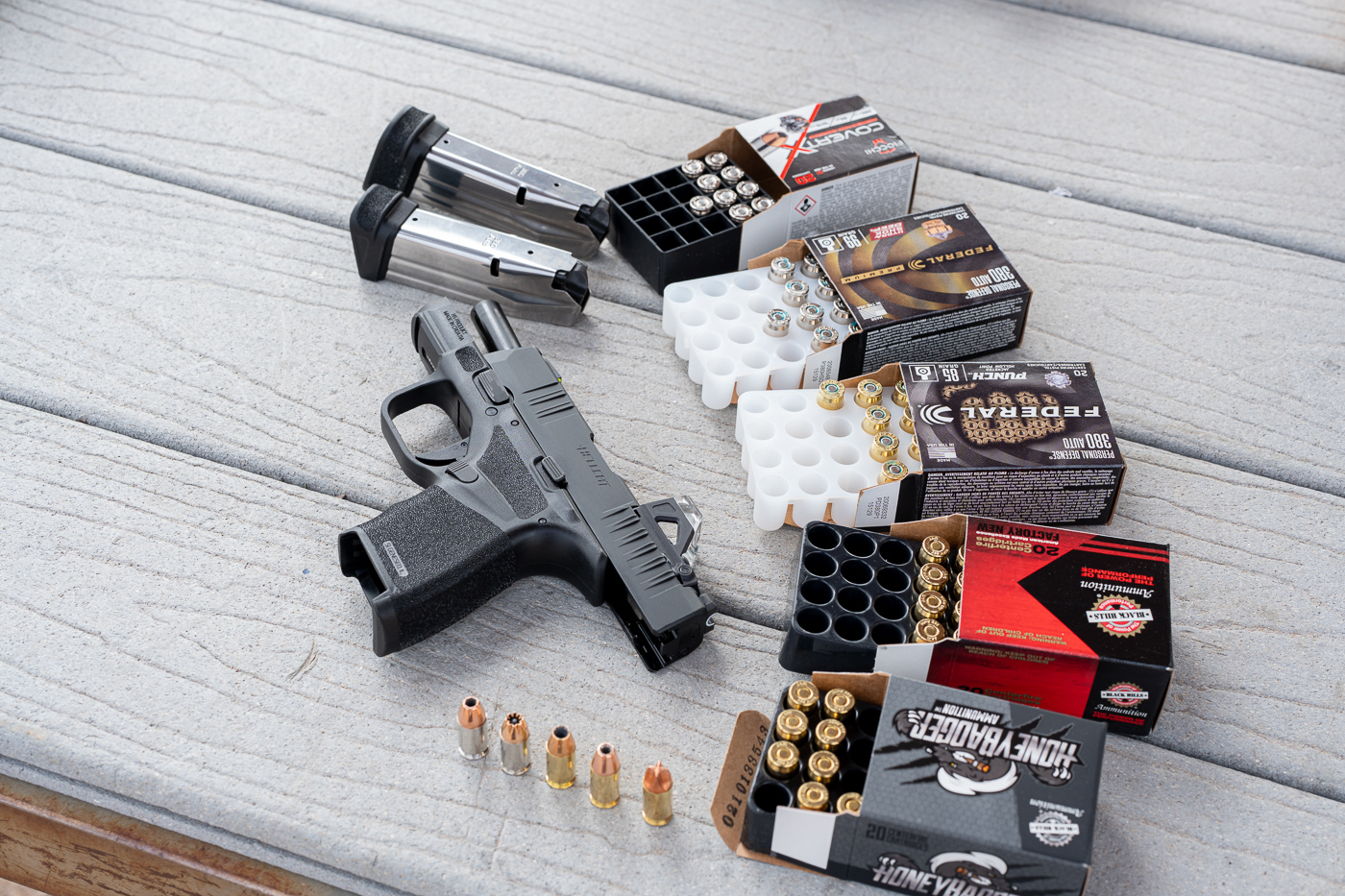 A collection of .380 Auto defensive loads photographed on a wooden range table with their packaging partially visible. The photo shows different hollow point designs and projectile finishes used in performance testing with the Springfield Armory Hellcat 380 ACP pistol. The boxes are arranged neatly to display the brands and grain weights for context on the test data. Some loose cartridges are positioned upright to show bullet cavity details and case color variations. The image gives readers a clear idea of what types of ammunition were used to assess feeding reliability, recoil, and accuracy. The natural outdoor lighting and neutral background reinforce the practical, range-based tone of the review. This image pairs well with sections covering ammo comparison and ballistic consistency for the Springfield Hellcat 380 ACP.
