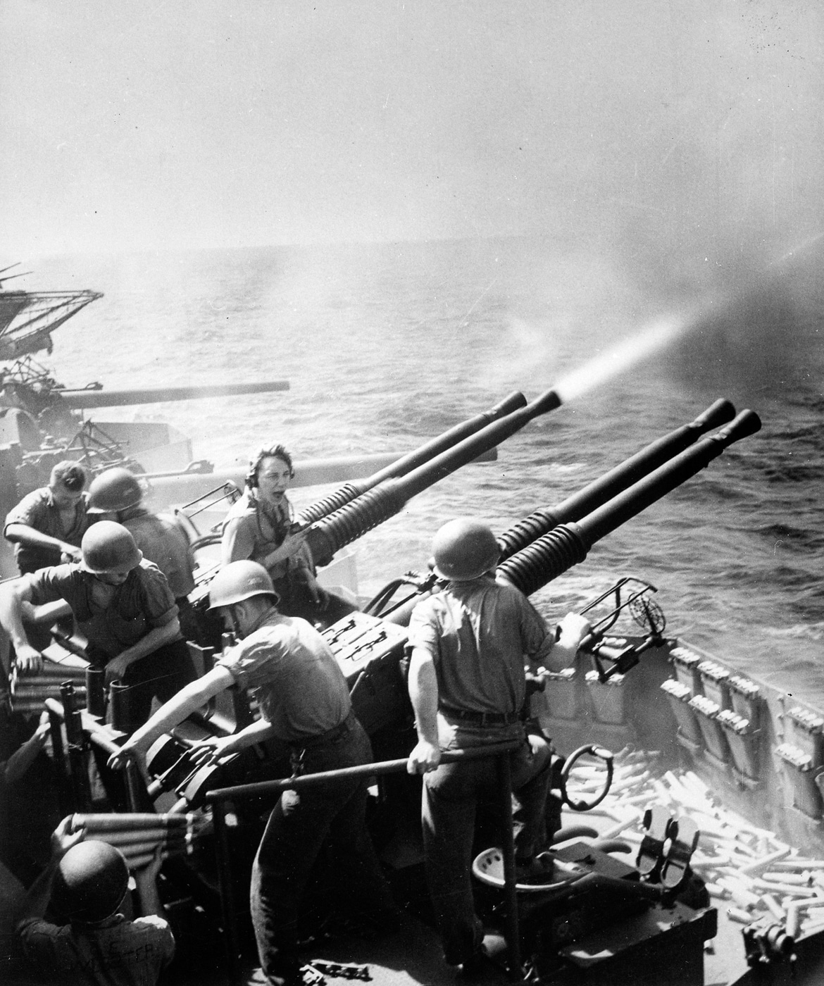 Black-and-white photograph of a 40mm Bofors anti-aircraft gun crew on the USS Hornet during World War II. Four sailors are visible—one at the sighting system, two loading four-round clips, and another standing ready with additional ammunition. The twin-barrel gun mount points skyward toward unseen aircraft as spent shell casings scatter across the deck. The ship’s railing and ocean horizon are visible behind the crew. The photo captures the tension of a kamikaze attack alert and the teamwork required to keep the Bofors firing. It illustrates how these Swedish-designed guns became the backbone of U.S. Navy air defense against Japanese Kamikazes in World War II.
