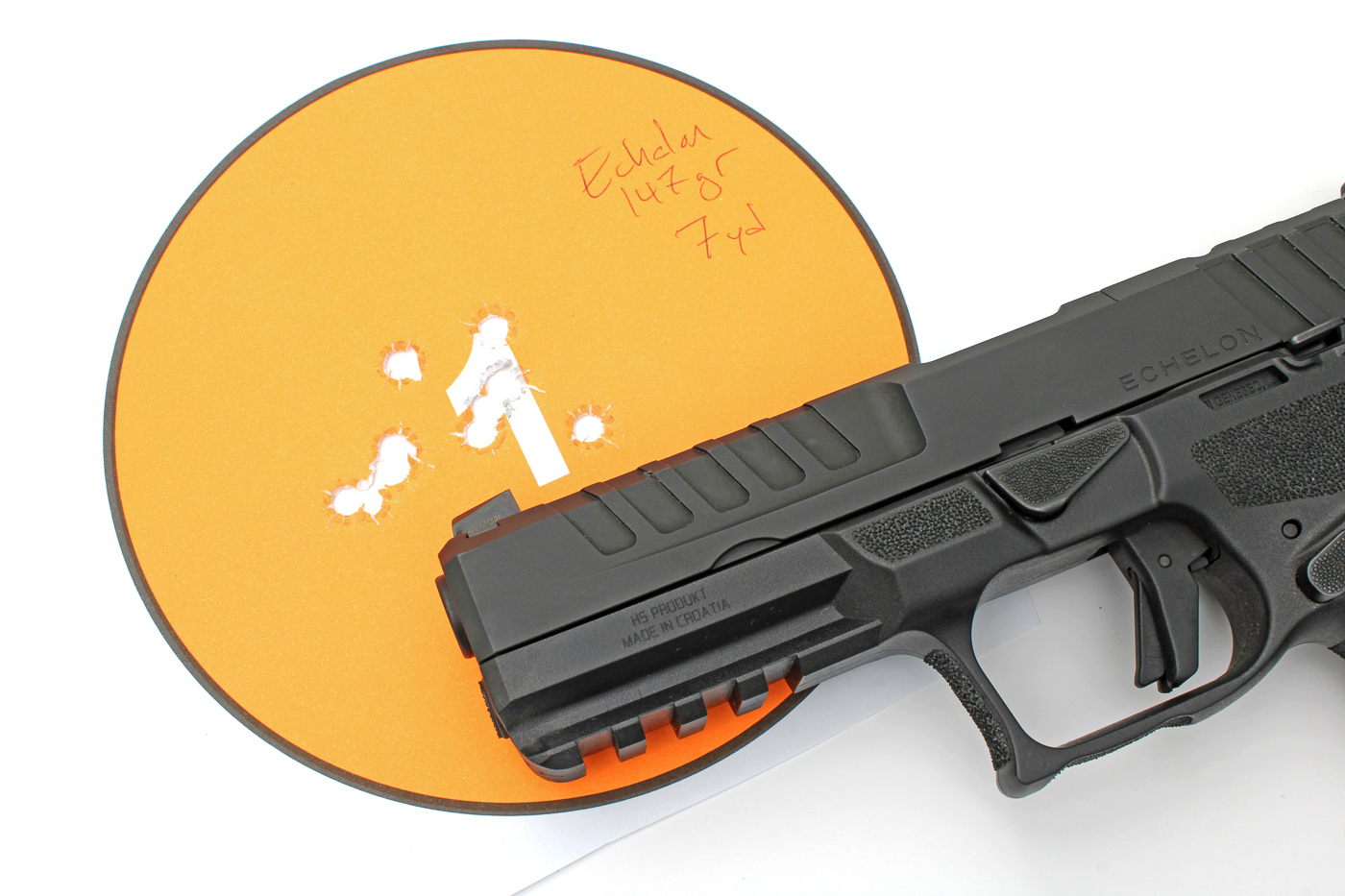 The photograph shows a California-compliant Springfield Echelon pistol placed next to a box or row of Remington UMC 147-grain FMJ 9mm cartridges and a paper target marked with tight groups. The handgun is displayed on a shooting bench, with the ammunition and shot target arranged for clarity. The target shows multiple holes grouped closely together, illustrating the pistol’s practical accuracy with this specific factory load. This image is intended to document benchrest accuracy results, supporting the article’s claims about performance and reliability of California-compliant Echelon pistols with common 9mm ammunition. The grip, slide, and controls of the pistol are visible, as are the cartridge casings and markings on the target.