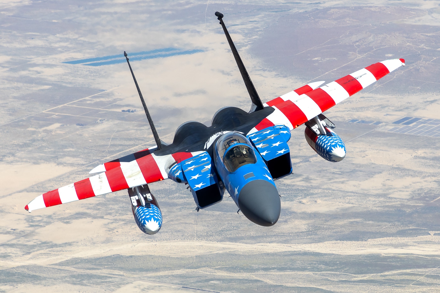 A California Air National Guard F-15C Eagle is shown with a distinctive American flag paint scheme, combining red and white stripes along the wings and fuselage with blue accents and white stars near the nose and intake areas. The jet retains its standard gray in other areas, with its tail flash number “113” visible. This aircraft, part of the 144th Fighter Wing, was given the patriotic artwork after logging more than 10,000 flight hours. The design celebrates both the service of the aircrew and the heritage of the Guard. The image captures the fighter in flight under bright conditions, highlighting the contrast between the painted motif and the structural details. The paint job does not degrade the aircraft’s operational markings, and the jet remains fully functional. The photo reflects how units sometimes commemorate achievement through symbolic liveries.