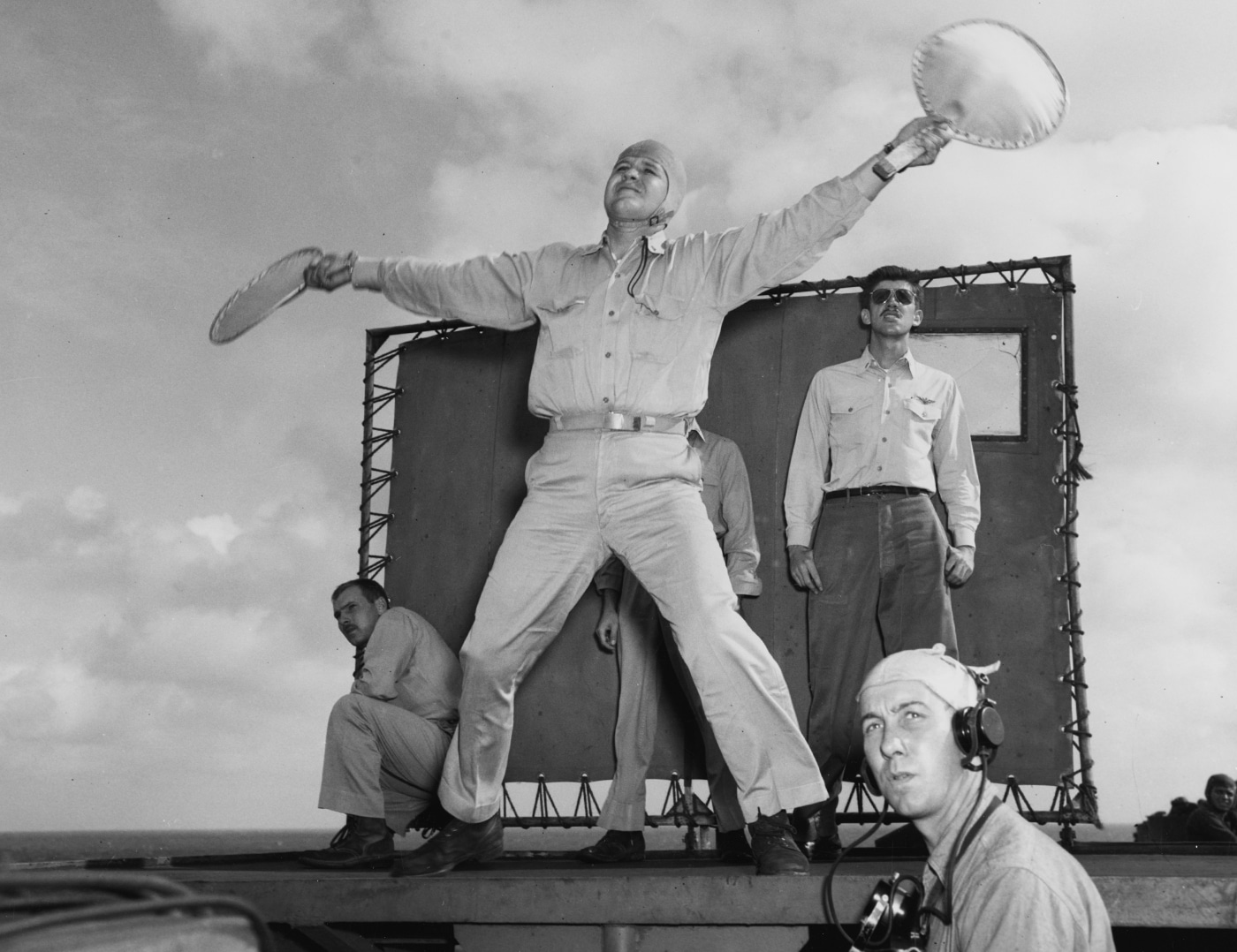 This photo features Ensign R. J. Grant serving as the landing signal officer (LSO) on USS Enterprise (CV-6) during active flight operations in March 1945. He stands near the stern of the flight deck, holding signaling paddles or flags, intently watching as an incoming aircraft lines up for landing. Wearing a helmet and flight deck gear, Grant uses precise hand and arm motions to guide pilots safely onto the moving deck. Other crew members and safety equipment are visible nearby, ready to assist in case of emergency. The ocean and wake of the carrier are visible in the background, highlighting the constant movement and challenge of carrier operations. The LSO’s position was exposed to noise, wind, and danger, underscoring the importance of his expertise and focus. The photo shows a critical moment in the daily routine of wartime flight operations, with Ensign Grant’s role essential to safe recoveries on USS Enterprise.