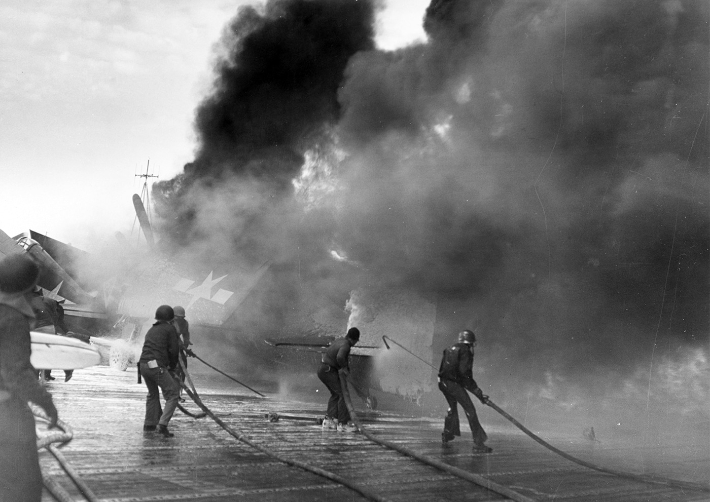 This photo shows sailors aboard USS Enterprise (CV-6) engaged in firefighting efforts after a kamikaze attack off Japan on March 20, 1945. Flames and thick black smoke billow from several burning F6F Hellcat fighter planes on the flight deck, which are visibly damaged and engulfed in fire. Crew members, equipped with hoses and protective gear, work in teams to spray water and foam onto the blaze, moving rapidly to prevent the fire from spreading to other aircraft or munitions. The deck is littered with debris from the explosion, and the chaotic scene reflects the intensity of damage control work in battle. Other sailors assist with moving undamaged aircraft and clearing pathways for firefighters. The urgency and danger of this moment are clear, showing how critical quick action was to saving the ship. This event was one of several kamikaze hits Enterprise survived during the final months of World War II.