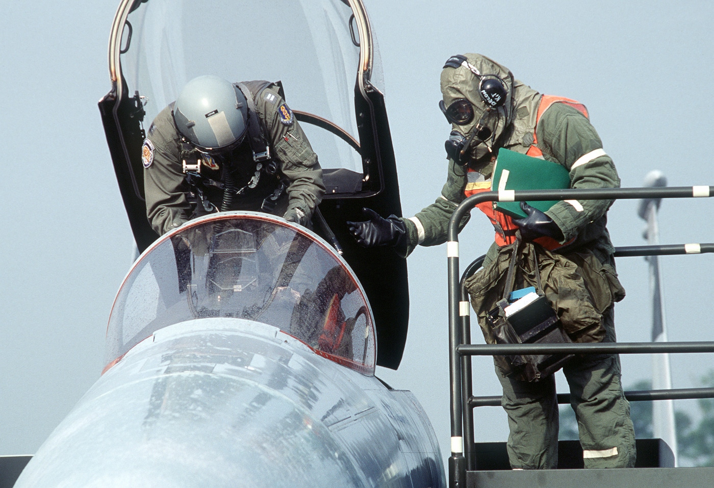 SSgt Richard Auclair of the 33rd Equipment Maintenance Squadron, dressed in full chemical protective gear, assists the pilot of a 58th Tactical Fighter Squadron F-15C Eagle during a Coronet Phaser deployment in Europe. The photo shows Auclair extending a hand to the pilot, who is climbing out of the cockpit after a flight. The protective MOPP (Mission Oriented Protective Posture) suit and mask reflect the chemical warfare training scenario taking place. Behind them, ground support equipment and the F-15’s twin tails are visible on the flight line. The Coronet Phaser program was designed to test how rapidly U.S.-based units could deploy and operate under combat-like conditions overseas. This image captures the cooperation between maintenance and flight crews during high-readiness exercises. It highlights both technical skill and adaptability in a simulated chemical threat environment.