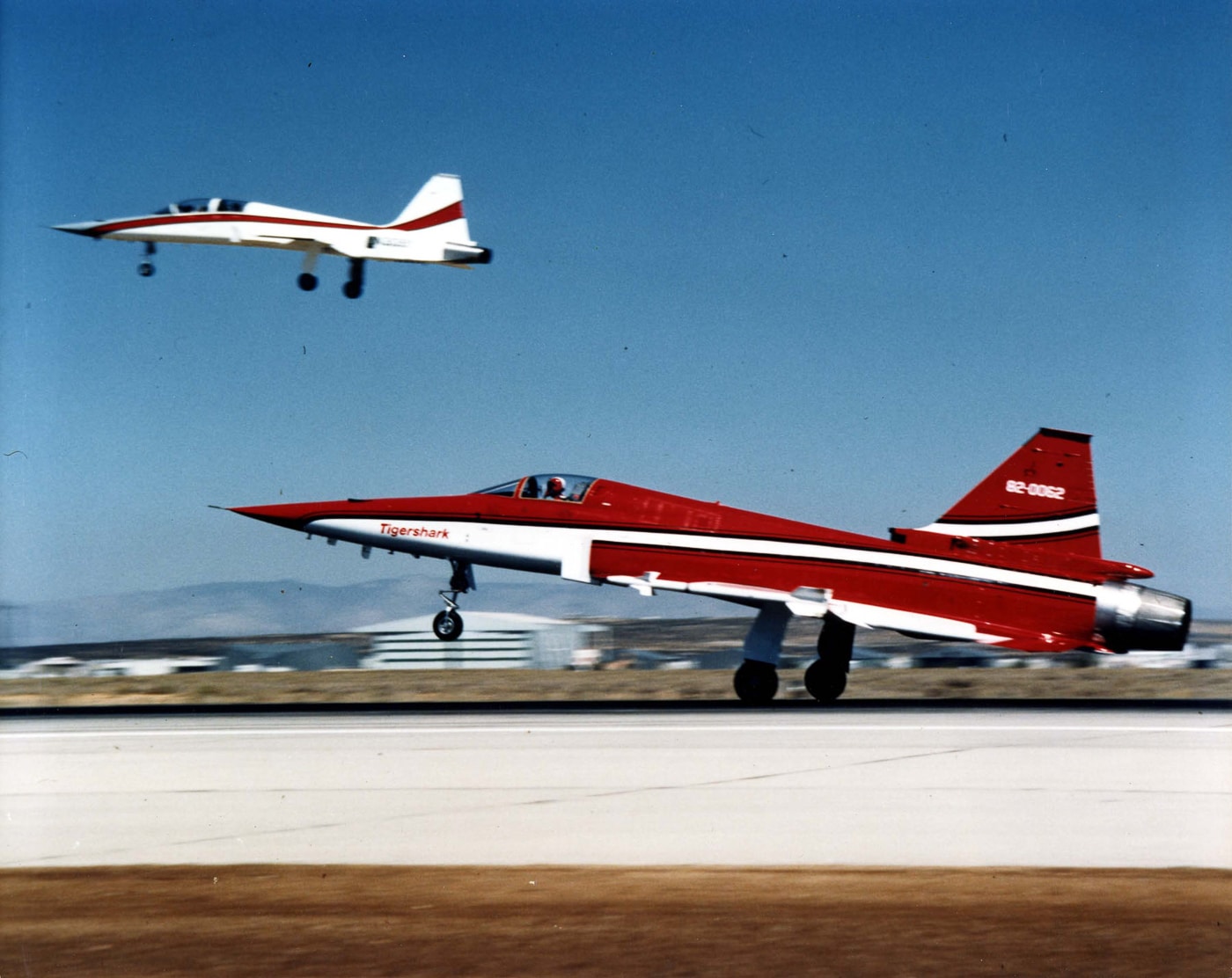 In this image, a Northrop F-20 Tigershark is shown flying in close formation with an F-5F Tiger II chase aircraft during a demonstration flight. The F-20 leads slightly ahead, showing its sleeker nose and modified airframe compared to the two-seat F-5F behind it. Both jets appear against a clear sky with light clouds, emphasizing their speed and precision flying. The chase plane was used to collect visual data and monitor the Tigershark’s flight behavior during testing. The aircraft markings are visible, reflecting Northrop’s continued refinement of its lightweight fighter line. This scene captures the direct lineage between the F-5 and F-20 programs. The overall setting conveys professionalism and engineering precision during evaluation.