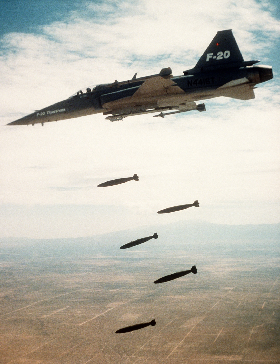 A Northrop F-20 Tigershark is seen mid-flight releasing several bombs during a demonstration over a weapons range. The photo captures the left side of the aircraft as the bombs fall away cleanly from its underwing pylons, trailing faint smoke against the clear blue sky. The jet’s gray fuselage and sharp aerodynamic lines are visible in bright sunlight. Northrop conducted this demonstration to test the fighter’s bomb-release mechanisms, accuracy, and flight control response under load. The aircraft remains perfectly level, indicating stable handling throughout the maneuver. The faint outline of terrain below adds scale and realism to the test environment. This image illustrates the F-20’s capability as a precise, multirole fighter platform.