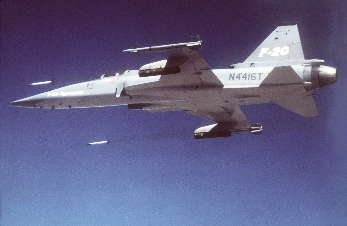 A Northrop F-20 Tigershark is captured midair firing a series of rockets from pods mounted beneath each wing. The rockets leave bright orange exhaust trails and plumes of white smoke as they streak toward a target area below. The aircraft maintains level flight, showing minimal movement or deviation from its trajectory. The gray paint of the F-20 contrasts sharply against the blue sky, while sunlight reflects along its fuselage. This demonstration was part of weapons integration testing to evaluate stability, accuracy, and control during rocket release. The scene showcases the fighter’s balanced aerodynamic design and steady flight characteristics. It conveys both power and precision during live ordnance evaluation.