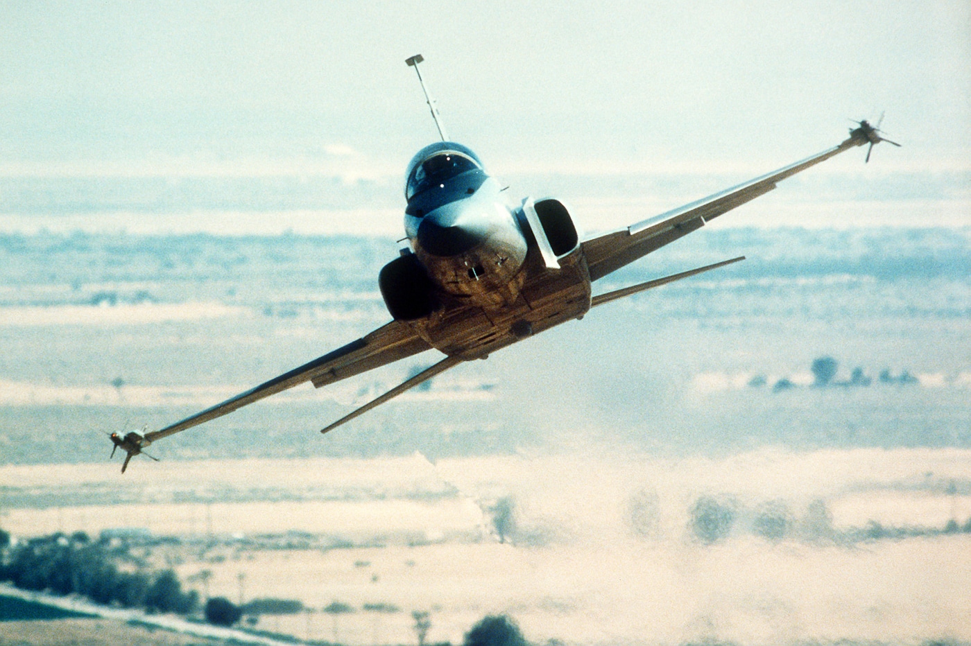 A Northrop F-20 Tigershark is captured performing a high-speed, low-altitude pass during a public demonstration at Edwards Air Force Base. The jet skims just above the runway, leaving heat shimmer in its wake as it races past at tremendous speed. The aircraft’s sleek gray fuselage and sharp nose are visible in profile, with the desert terrain and hangars of Edwards AFB in the background. Dust can be seen trailing slightly behind the aircraft, emphasizing its proximity to the ground. This maneuver was part of Northrop’s demonstration program to showcase the F-20’s stability and control authority at low altitude. The image captures the power, precision, and refined handling that characterized the Tigershark’s flight performance. The lighting and background give a clear sense of realism and technical display.