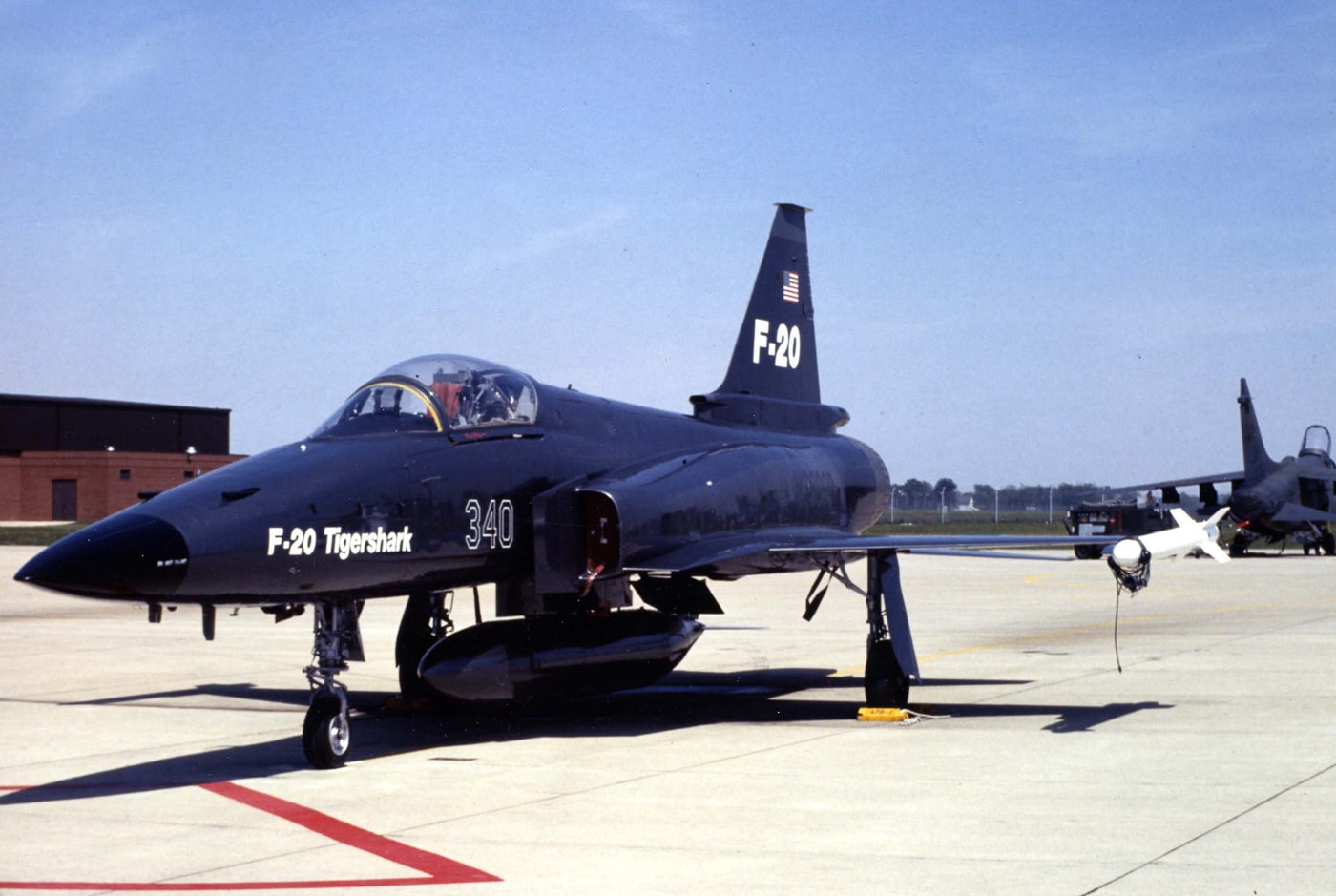 A Northrop F-20 Tigershark is parked on the flight line wearing a U.S. Air Force Aggressor paint scheme. The aircraft’s camouflage features gray and blue tones designed to resemble Soviet-style fighters used in dissimilar air combat training. The Tigershark’s clean, streamlined fuselage, single-engine layout, and sharp nose are clearly visible. Its canopy is closed, and the aircraft rests under daylight with ground equipment nearby. This version of the F-20 was tested for potential Air National Guard and Aggressor training roles. The paint scheme emphasized its intended use as a realistic adversary aircraft for pilot training.