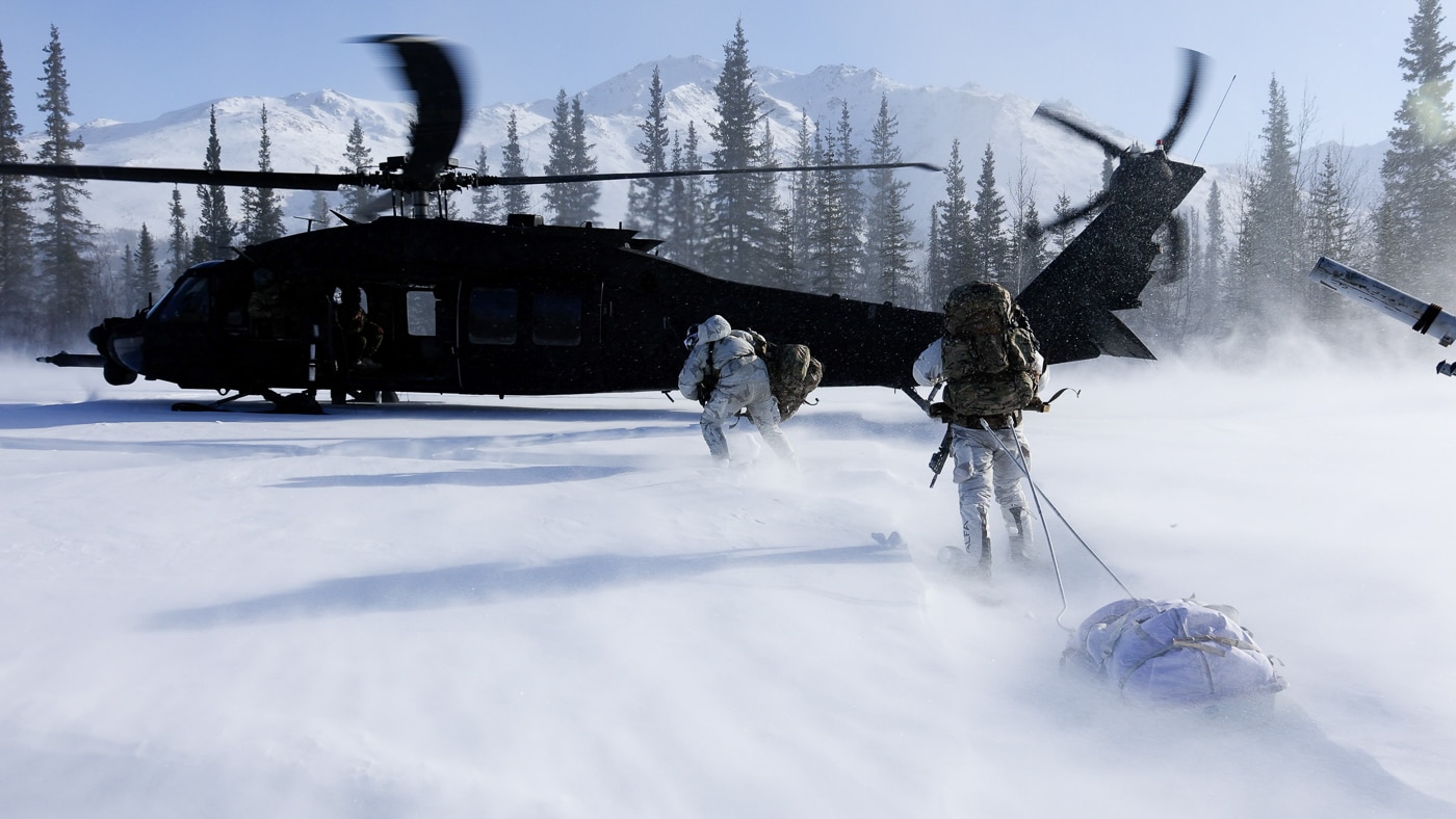 The image shows U.S. Army Green Berets from the 10th Special Forces Group (Airborne) walking across a snow-covered landscape toward an MH-60 Black Hawk helicopter assigned to the 160th Special Operations Aviation Regiment (Airborne). The helicopter is on the ground with its rotors still or slowly turning, positioned in a remote area near Wiseman, Alaska, during Exercise ARCTIC EDGE 2022. The Green Berets are wearing heavy cold-weather gear, including parkas, gloves, and face coverings, carrying rucksacks and weapons suited for arctic operations. Snow, ice, and rugged terrain dominate the scene, with mountains or forest visible in the distance. The exercise emphasizes cold-weather mobility, teamwork, and rapid insertion in harsh environments. This joint operation demonstrates how the 160th SOAR supports Army special operations in some of the world’s most challenging climates. The photo highlights the adaptability and resilience required for success in Arctic missions.