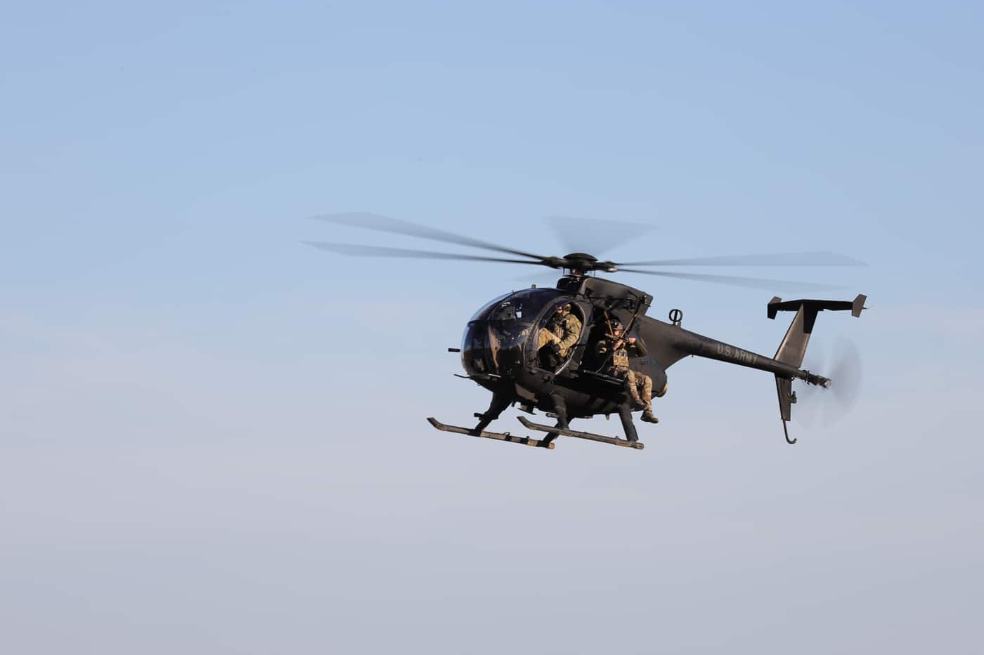 The image features Green Berets of the 5th Special Forces Group conducting sniper training from an MH-6 Little Bird helicopter, operated by the 160th Special Operations Aviation Regiment. The MH-6 is hovering or in slow flight, doors open, with at least one sniper in full tactical gear positioned on the skids or inside the aircraft, aiming a precision rifle at a ground target. The terrain below may be an open training range, with visible targets, berms, or other range features. The scene emphasizes the close integration between Army Special Forces and the Night Stalkers, highlighting the unique challenges of aerial marksmanship. Safety harnesses, communication headsets, and other specialized equipment are evident. The compact size of the Little Bird and the skill required to maintain a stable firing platform are central to the photo’s context. This type of training is vital for developing rapid-response sniper teams for special operations missions.