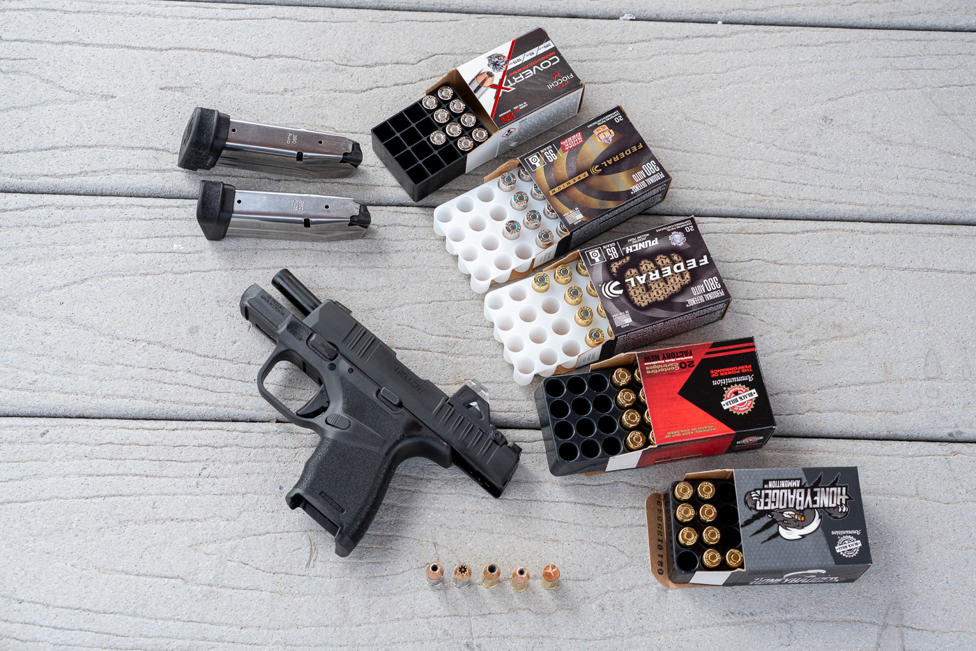 A Springfield Hellcat 380 ACP pistol rests on a shooting-bench surface at an outdoor range, positioned next to several boxes and loose cartridges of .380 ACP self-defense ammunition. The photo shows jacketed hollow points, bonded bullets, and defensive load boxes arranged to indicate the different rounds used during testing. The pistol is pointed downrange and the shooter’s protective gear is visible in the background, establishing a controlled range environment. This composition emphasizes that the review included live-fire testing for reliability, accuracy, and velocity. Visible details include the Hellcat’s short slide, textured grip, and magazine baseplates used during the session. The image is useful for illustrating sections about ammo selection, terminal performance, and what defensive loads function best in a micro-compact platform. It supports the article’s comparison of different .380 Auto defensive options with the Springfield Hellcat 380 ACP.