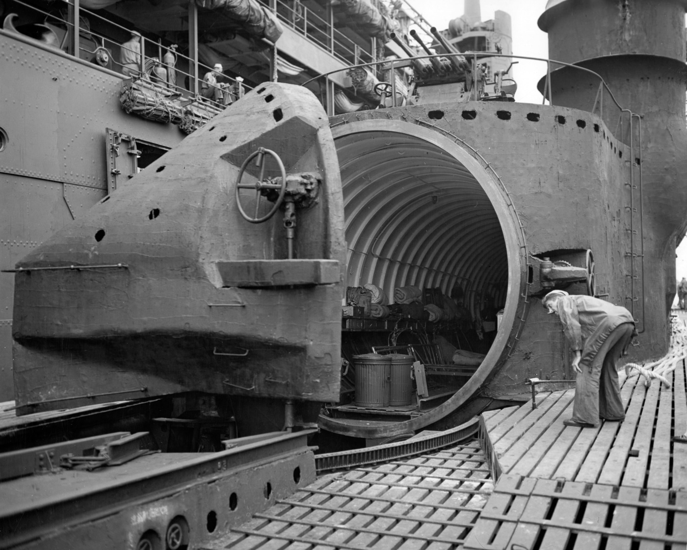 A black-and-white photograph showing U.S. Navy personnel examining the watertight aircraft hangar on the deck of the Japanese I-400 submarine shortly after its capture. The image reveals the large cylindrical hangar structure positioned forward of the conning tower, where the Aichi M6A Seiran floatplanes were stored with their wings and tail sections folded. The I-400 class submarines were capable of carrying two or three aircraft, which could be assembled and catapult-launched within minutes. This inspection photo highlights the innovative design and impressive size of Japan’s underwater aircraft carriers, which combined aviation and naval technology in a single platform.