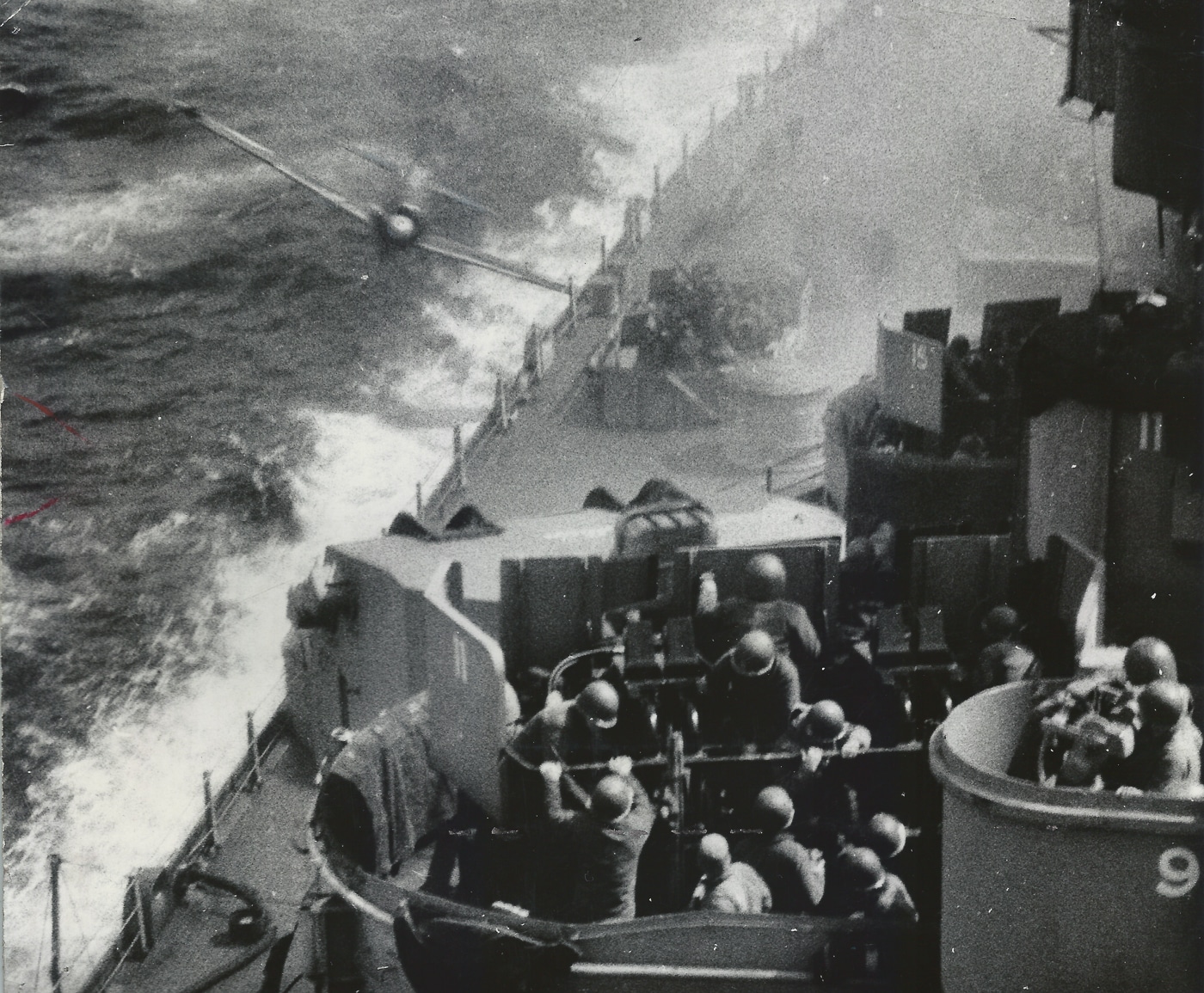 A historical black-and-white photo showing a Japanese kamikaze plane crashing into the side of the USS Missouri, an Iowa-class battleship, during World War II. Smoke and debris fill the air as crew members rush to contain the damage. The photo captures the intensity of naval combat in the Pacific and the destructive nature of suicide attacks. The ship’s massive guns and armored hull remain intact despite the explosion. U.S. sailors can be seen on deck responding to the aftermath, illustrating the resilience of the American fleet under kamikaze assault.