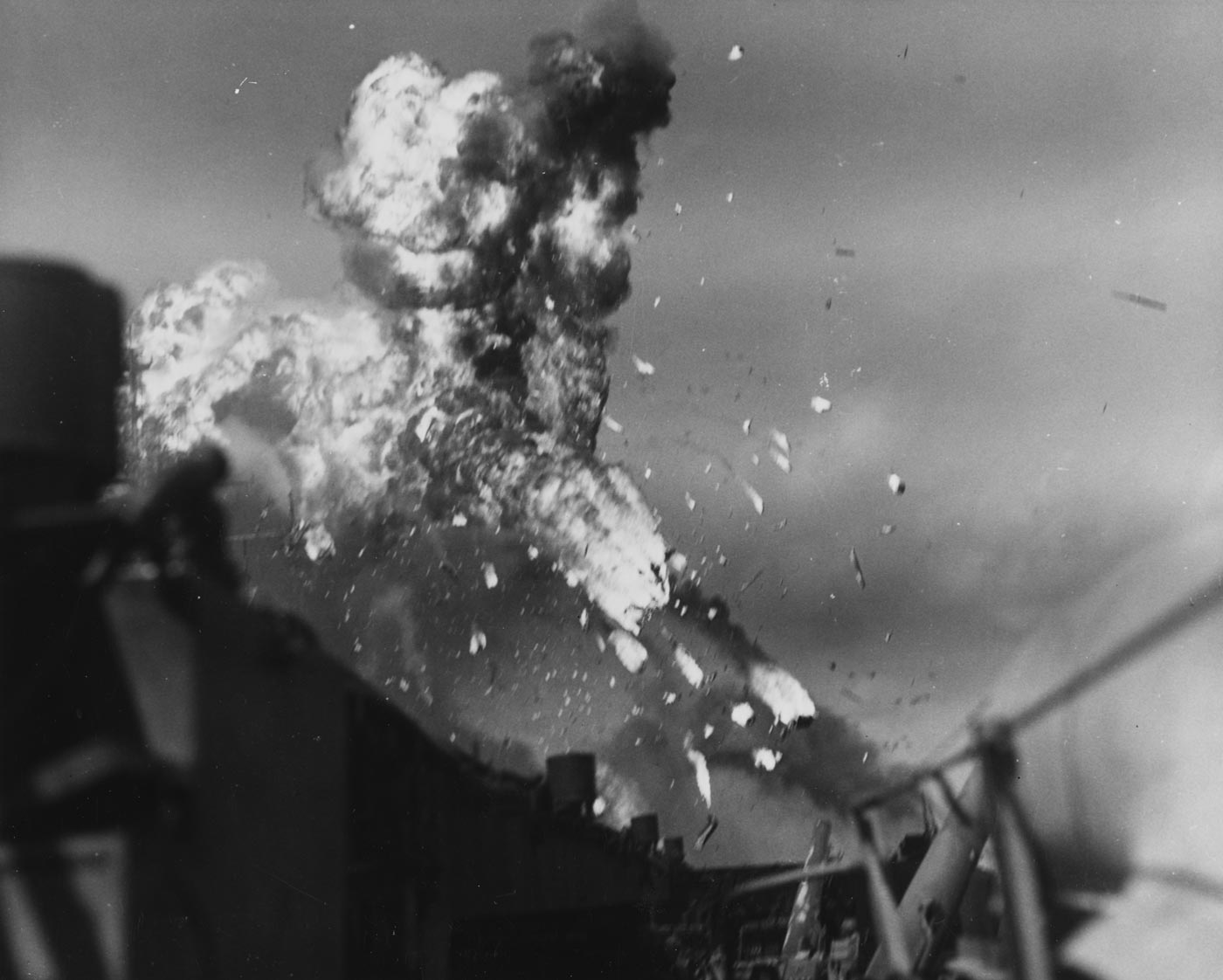 A dramatic wartime photograph shows the USS Intrepid (CV-11) engulfed in fire and smoke moments after being struck by a Japanese kamikaze plane on November 25, 1944. The explosion sends flames and fragments high above the flight deck, where several aircraft are visible amid wreckage. Crewmen can be seen running toward the flames carrying hoses and extinguishers as black smoke pours into the sky. The photo captures the chaos and violence of kamikaze attacks that plagued U.S. carriers during the Philippine operations. Despite the damage, the Intrepid remained afloat and operational within hours. This image powerfully illustrates the destructive force of Japanese Kamikazes in World War II and the resilience of U.S. Navy crews under fire.