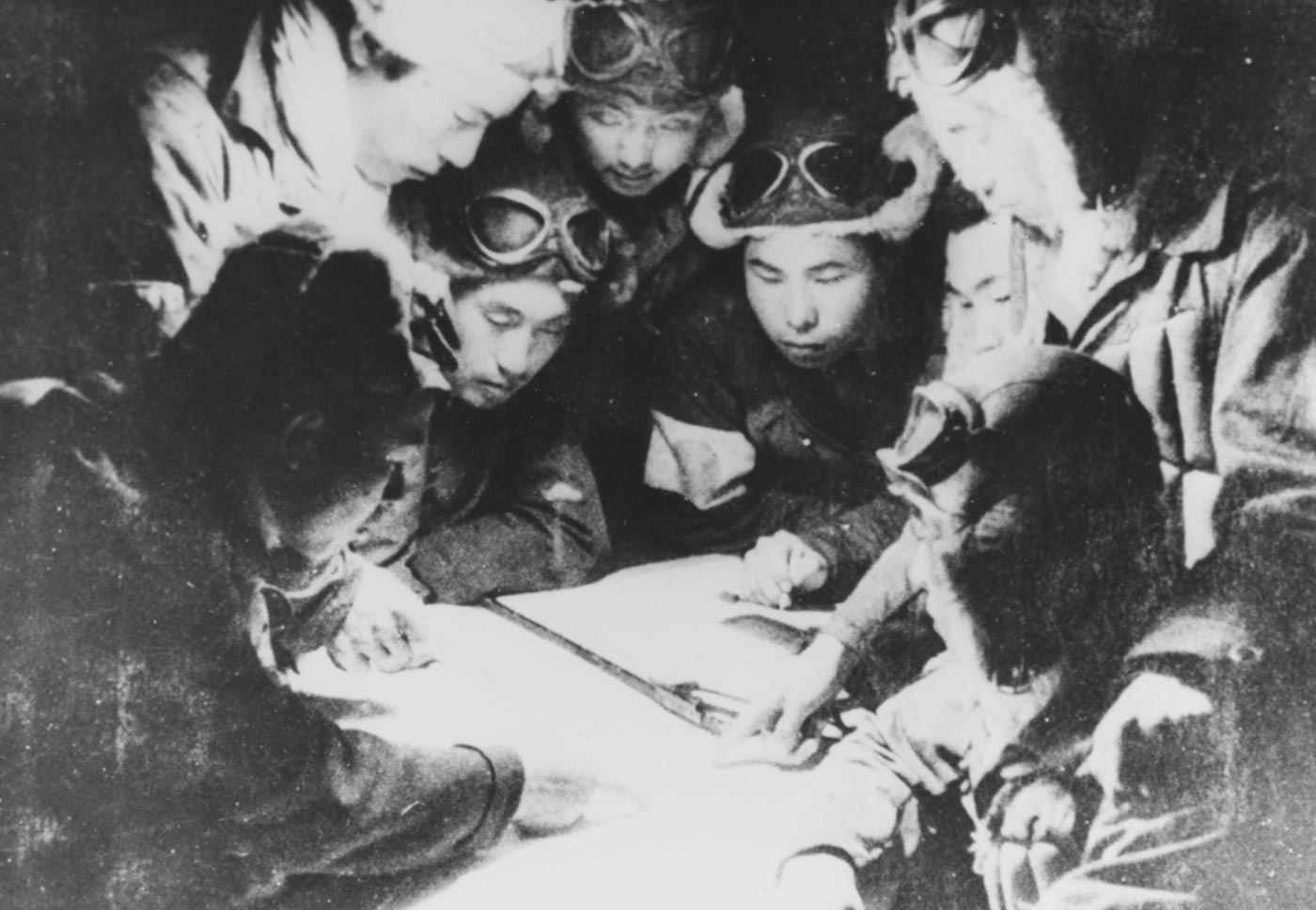 A black-and-white photo showing a group of young Japanese kamikaze pilots sitting on the floor of a briefing room the night before their mission in World War II. An officer stands at the front beside a chalkboard or map, gesturing as he outlines flight routes and targets. The men wear flight suits or light uniforms, their expressions focused and subdued. Oil lamps or candles cast a soft glow, emphasizing the intimacy of the setting. Papers, maps, and headbands bearing the rising sun emblem lie scattered nearby. The image conveys the human side of Japan’s kamikaze program — the mixture of duty, fatalism, and ritual that defined these final preparations. It provides an emotional glimpse into the mindset of Japanese Kamikazes in World War II.