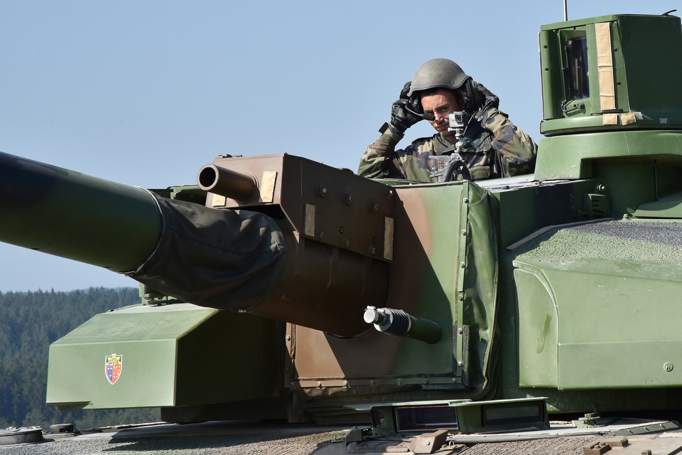 The photo shows a French Leclerc tank commander standing in the open turret hatch during a training exercise. The commander is wearing standard military gear, including a helmet and communications headset. This elevated position provides enhanced visibility over the battlefield compared to viewing through periscopes. The commander is responsible for giving orders to the gunner and driver, coordinating movement, and identifying targets. The Leclerc’s panoramic sighting system and digital battle management tools support this role, but direct observation remains crucial. The image highlights the leadership and situational awareness required of a tank commander during training.