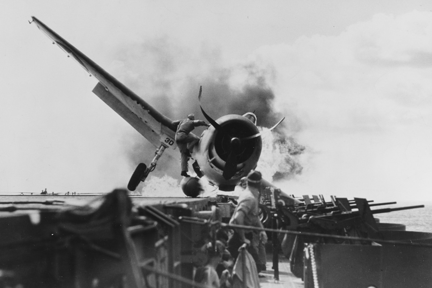 This powerful photo shows Lt. Walter Chewning, the assistant flight deck officer on USS Enterprise (CV-6), as he climbs onto the wing of a burning F6F Hellcat fighter. Flames and smoke engulf part of the aircraft after a crash landing, while Chewning acts quickly to reach the trapped pilot in the cockpit. Wearing flight deck gear and helmet, he braces himself against the intense heat to pull the canopy open. Other crew members are seen nearby, rushing with firefighting equipment and ready to assist. The urgency and danger of the scene are clear, with the fire threatening both the pilot’s life and the safety of the ship. This moment captures the bravery and quick decision-making required during emergencies on a carrier flight deck. The actions of Chewning and his team helped save lives in the high-risk environment of World War II naval aviation.
