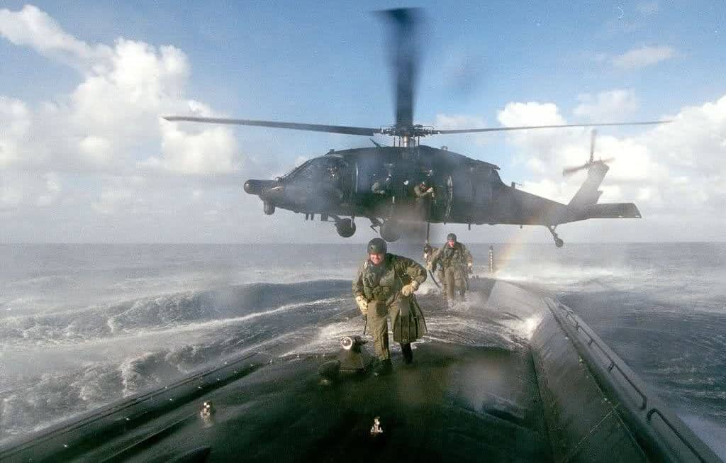 The image captures an MH-60L Black Hawk helicopter from the 160th Special Operations Aviation Regiment (Airborne) hovering just above the deck of a surfaced submarine, delivering a team of soldiers from the 7th Special Forces Group. The soldiers, equipped with tactical gear, helmets, and possibly scuba or maritime equipment, are seen exiting the helicopter onto the submarine’s narrow deck. The helicopter’s rotors are spinning, and the aircraft is positioned with precision to allow safe and rapid disembarkation. The submarine is partially visible in open water, with waves and the horizon in the background. This scene highlights the complexity and risk involved in maritime special operations, emphasizing the close cooperation between Army aviation and Special Forces. The photo showcases the technical skill of the Night Stalkers pilots and the readiness of the 7th SFG team. The operation demonstrates the unique insertion techniques used for clandestine maritime missions.