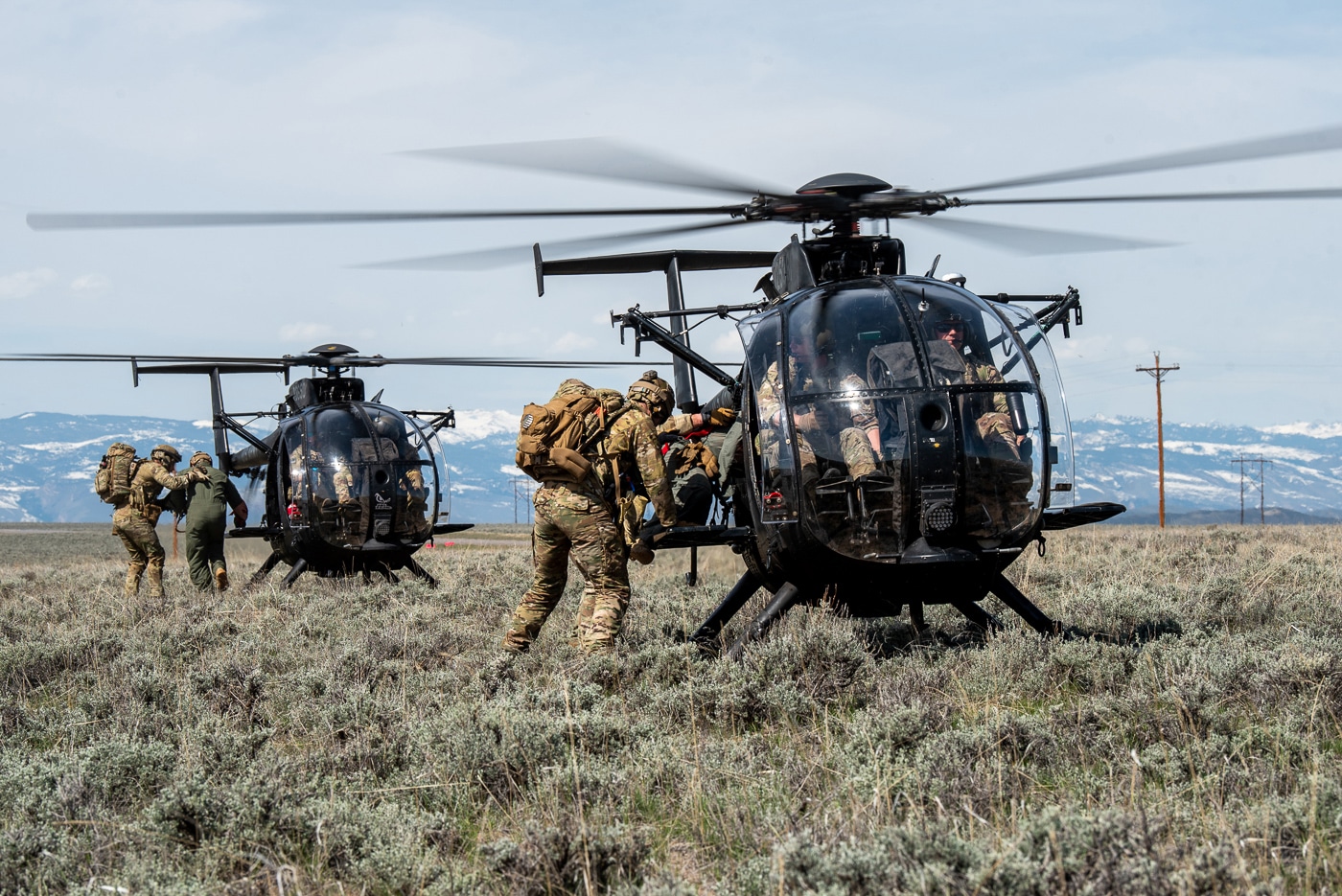 The image shows MH-6M Little Bird helicopters from the 160th Special Operations Aviation Regiment (Airborne) landing in a remote or improvised field landing zone to extract U.S. Air Force airmen from the 123rd Special Tactics Squadron during Exercise Agile Chariot. The helicopters are small, agile, and configured for special operations, with open doors or external seating visible. Airmen, wearing tactical gear, helmets, and carrying rucksacks or weapons, are either boarding the helicopters or running toward them for rapid extraction. The environment is a grassy or dirt field, possibly with trees or limited cover in the background. The scene highlights the close coordination and fast-paced nature of special operations extractions, with pilots maintaining a low hover for quick embarkation. The exercise underscores the ability of the 160th SOAR to operate in austere environments and support joint missions with Air Force special tactics teams.