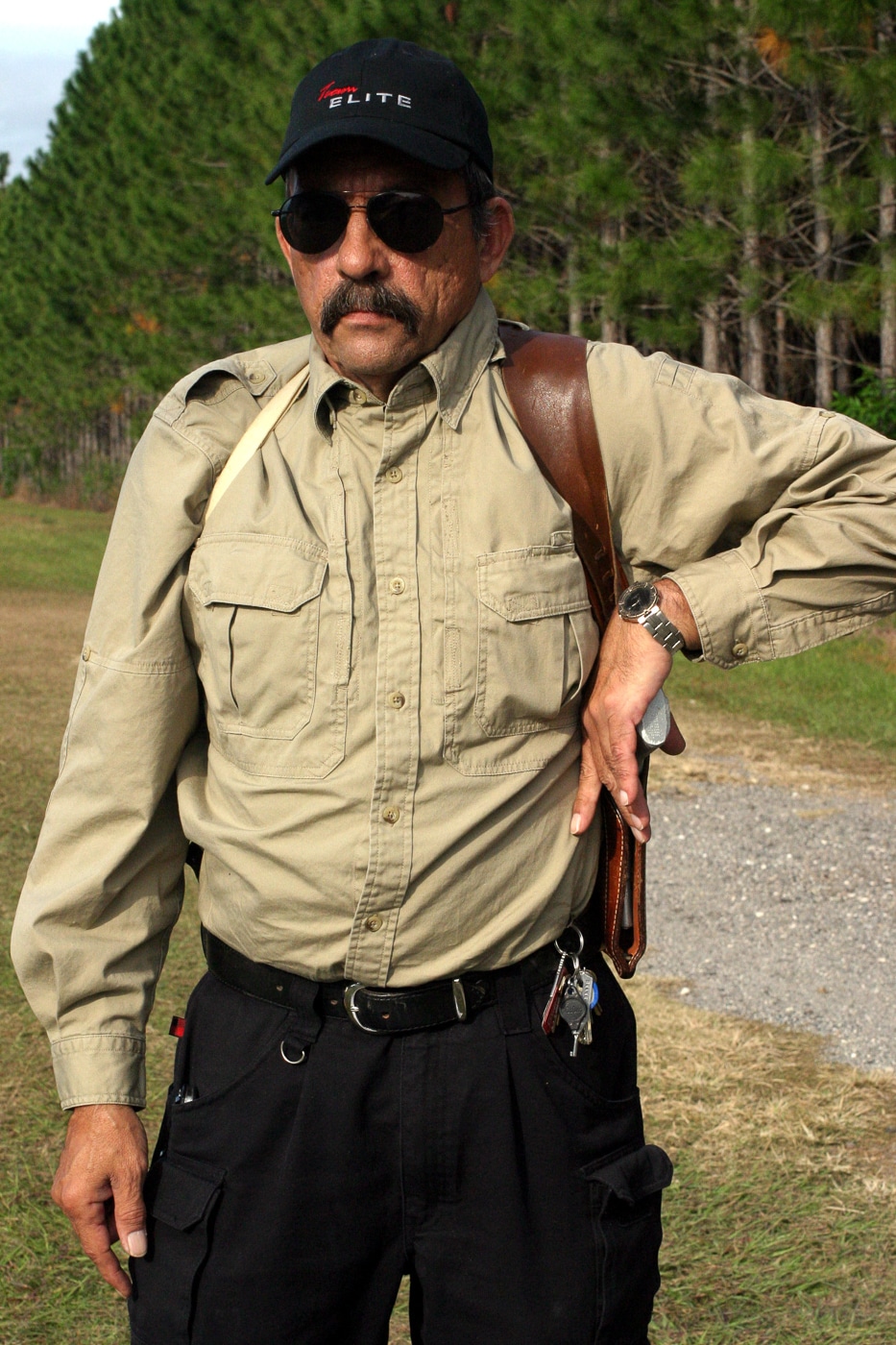 The image shows a man demonstrating a weak-hand draw from a shoulder holster, often referred to as a Fudd holster. His non-dominant hand reaches across his body to grip the handgun under his arm. The firearm is still partially holstered, and his trigger finger remains outside the trigger guard, showing proper safety technique. The shoulder rig’s straps are visible across his chest and shoulders, evenly distributing the weight of the pistol. The background is simple, allowing focus on the form and mechanics of the motion. The photo illustrates how the shoulder holster can provide ambidextrous access when the strong hand is unavailable. It supports the article’s broader point about practicality and adaptability in concealed carry methods.