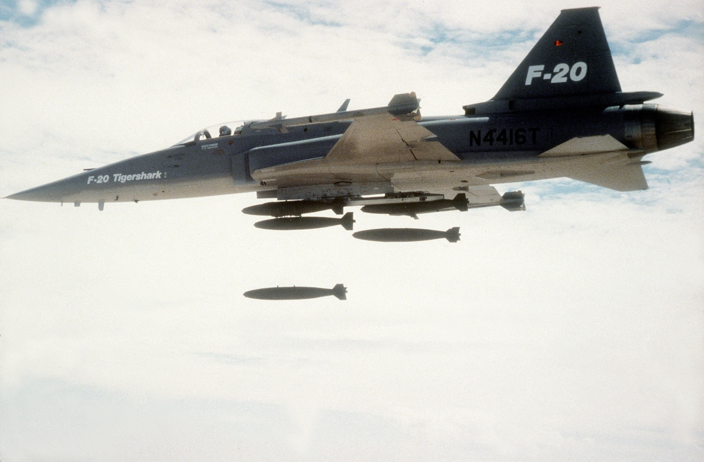 A left side, air-to-air view of a Northrop F-20 Tigershark shows the aircraft in level flight as it releases multiple practice bombs over a designated test range. The bombs can be seen falling in sequence beneath the aircraft, trailing faint smoke as they descend. The F-20’s streamlined fuselage, swept wings, and single-engine tail section are clearly visible in bright sunlight. The aircraft’s markings and gray camouflage contrast against the blue sky and the earth far below. This demonstration was part of weapons evaluation testing to confirm accuracy and stability during ordnance release. The photo captures both the precision and performance of the Tigershark during its development program. The image illustrates how the lightweight fighter handled air-to-ground strike missions with ease.