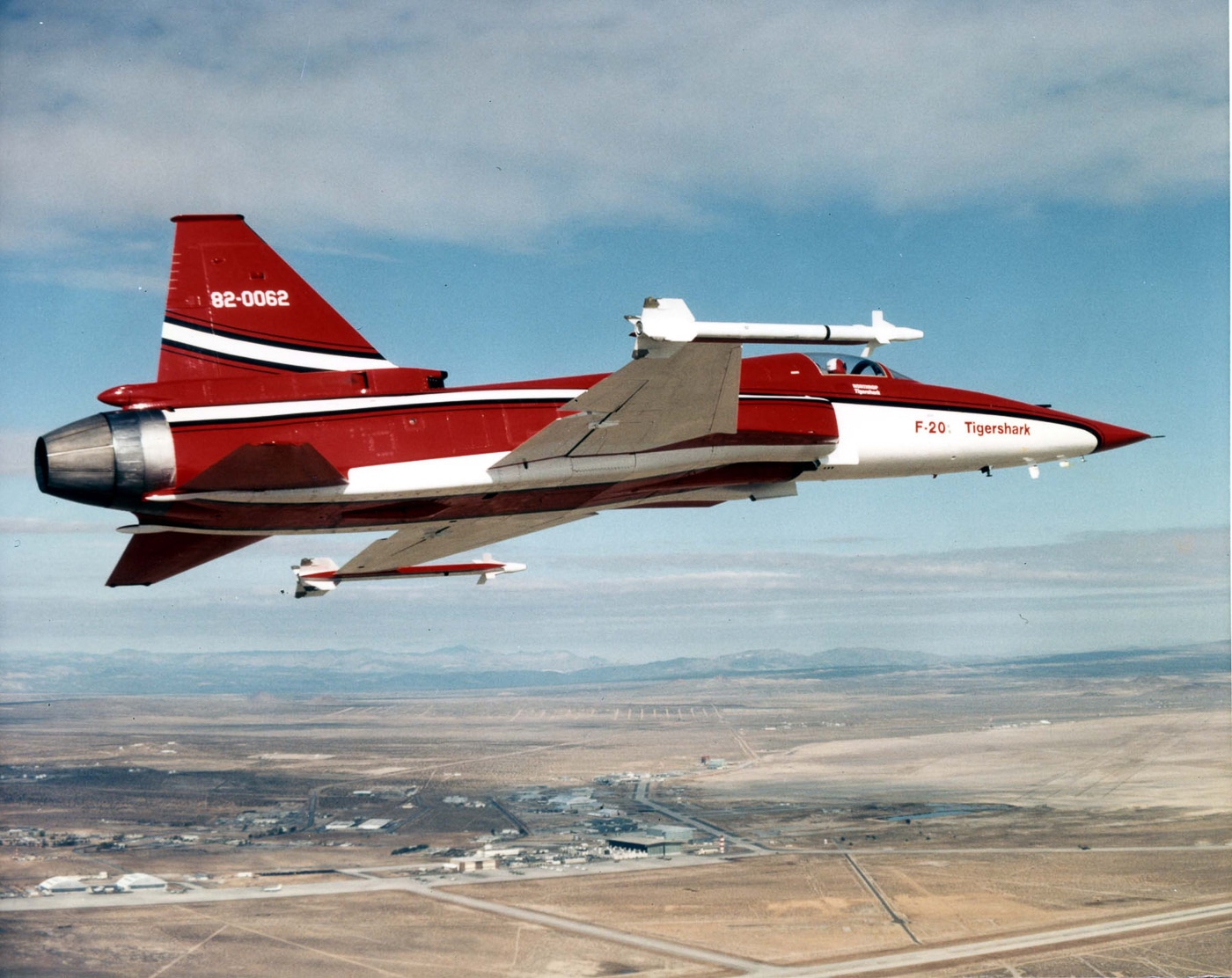 A Northrop F-20 Tigershark is captured midair during a test flight, banking gently under clear skies. The aircraft’s sharp nose, single air intake, and slender fuselage define its clean aerodynamic design. Its gray paint scheme contrasts with the blue sky, and sunlight highlights the aircraft’s control surfaces and canopy reflections. Northrop engineers conducted these flights to measure climb rate, stability, and acceleration under various power settings. The F-20’s test program confirmed its ability to reach Mach 2 speeds and rapid response readiness. The photo reflects the precision and technical focus of the Tigershark’s development phase. The background shows faint terrain below, emphasizing altitude and scale.
