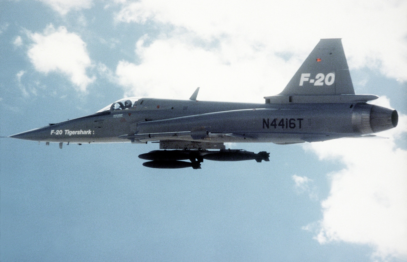A Northrop F-20 Tigershark is shown parked on the flight line with multiple practice bombs mounted on its underwing pylons and fuselage stations. The photo clearly shows the aircraft’s mid-mounted wings, single engine intake, and populated bomb racks, indicating a ground-attack loadout. The canopy is closed and the jet is pointed down the runway as if preparing for taxi or takeoff. Ground crew equipment and a tarmac surface are visible around the aircraft, giving context to pre-mission preparations. This view emphasizes the F-20’s ordnance-carrying capability and role as a light attack/strike platform. The description is useful for accessibility and for users searching keywords like F-20 payload, bomb racks, and air-to-ground configuration. The overall scene communicates readiness and the practical weapons integration of the Tigershark.