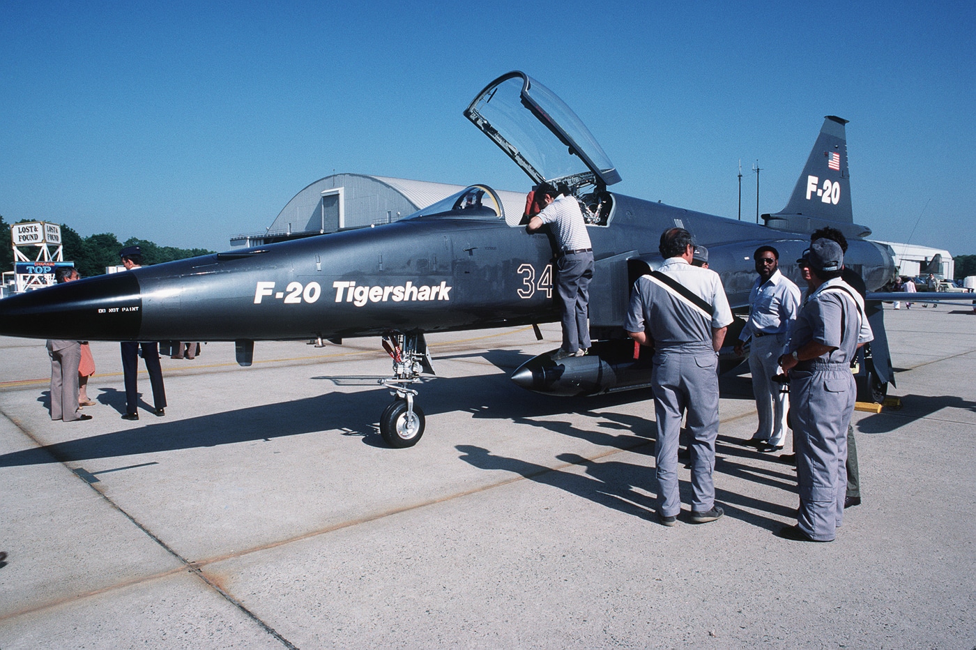 A Northrop F-20 Tigershark sits parked on the tarmac during an air show as members of the Northrop team stand nearby answering questions from spectators. The aircraft’s gray fuselage, single tail, and compact design are clearly visible under bright daylight. Visitors in casual clothing gather around the jet, taking photos and listening to explanations about its performance, avionics, and role as a lightweight fighter. The scene reflects a public outreach event where engineers and marketing representatives engaged directly with the crowd. Background elements include portable barriers, display signs, and nearby aircraft from other exhibitors. The F-20’s canopy is closed, and the aircraft appears in a clean, unarmed configuration. This image captures the human connection between manufacturer and public during the Tigershark’s promotional efforts.