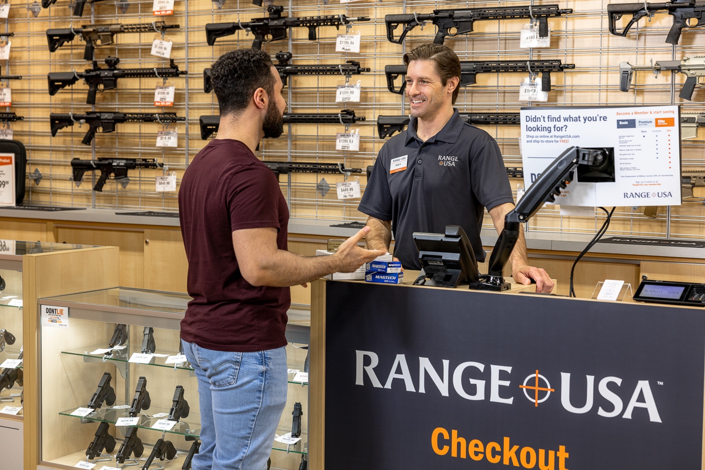 Photo of a Range USA employee speaking with a customer inside the retail section of a shooting range facility. The staff member is wearing a branded shirt and name tag, smiling while explaining product options or range procedures. The customer appears attentive and relaxed, standing near a display of firearms or accessories. The store interior is brightly lit and well-organized, with shelves of shooting gear and signage visible in the background. The image conveys Range USA’s focus on customer service, education, and safety. It illustrates the company’s commitment to creating a welcoming, informative experience for both new and experienced shooters.