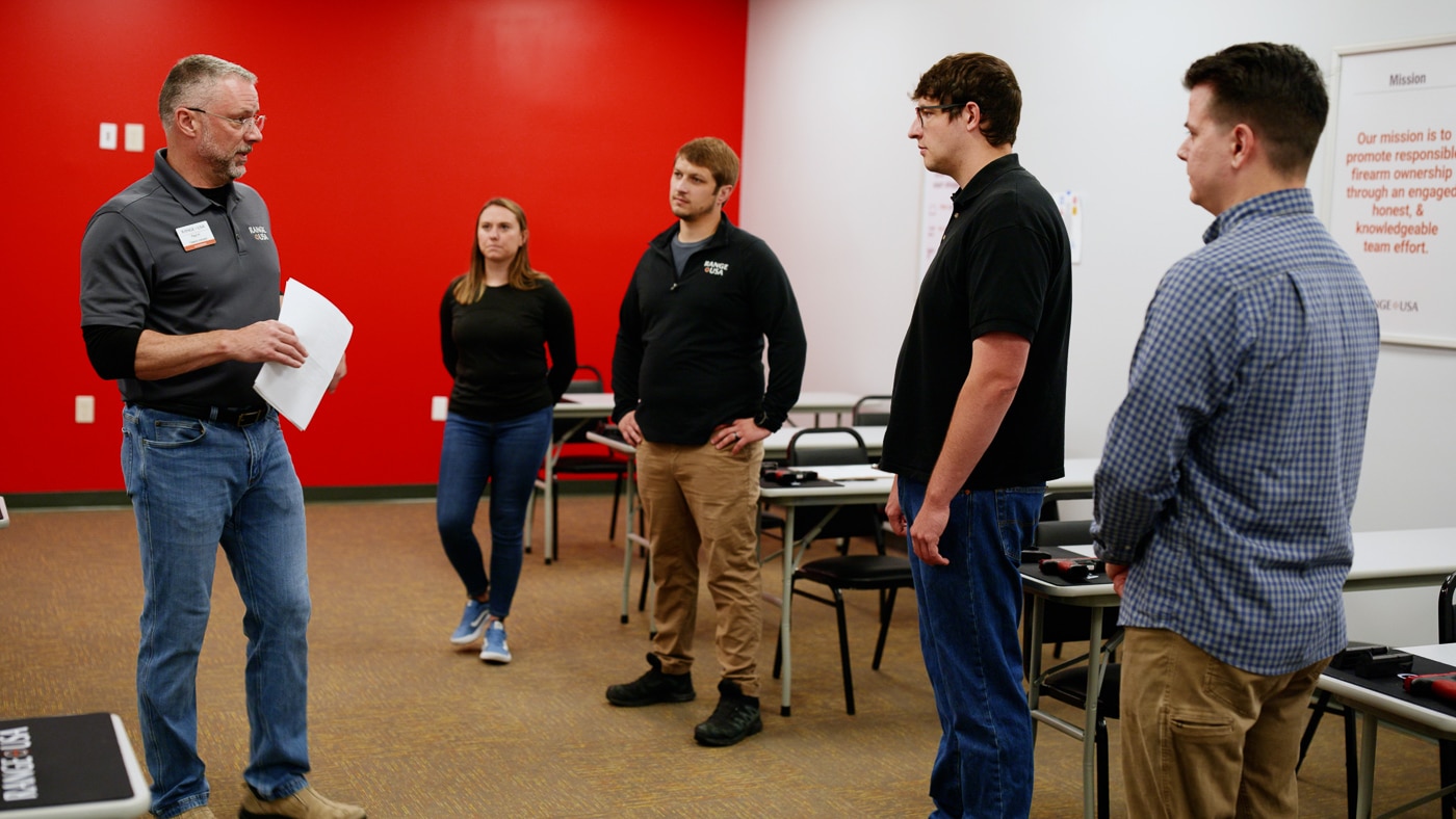 Photo of a Range USA firearms instructor leading a class for new shooters inside a bright, organized training room. Several participants sit at tables or stand near the range area while the instructor demonstrates firearm safety techniques. Visual aids and firearms training materials are visible on nearby tables. The group includes a mix of men and women, suggesting a welcoming environment for all experience levels. The image highlights Range USA’s focus on education, hands-on learning, and safety. It represents the company’s structured training programs that help new shooters gain confidence and foundational skills.