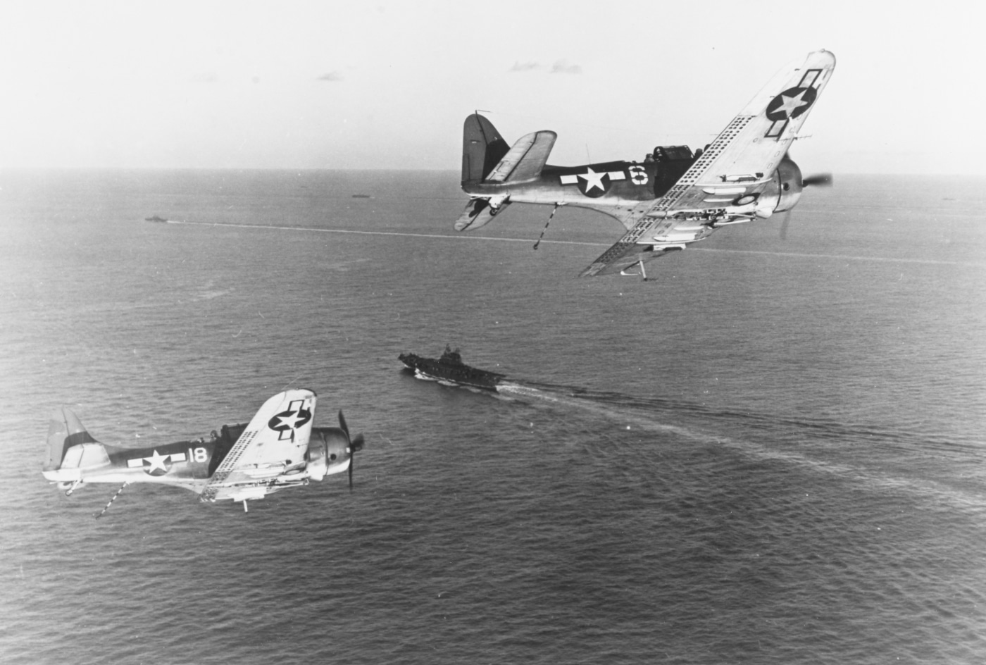 This photo shows a group of SBD Dauntless dive bombers maneuvering in the air as they prepare to land on the USS Enterprise (CV-6) following a combat strike during the Battle of Peleliu. The aircraft are flying in formation, wings level, as they make their final approach to the carrier’s flight deck. The ship is visible below, with crew members stationed and ready to assist with landings. The sea is calm, providing clear conditions for recovery operations. The dive bombers’ distinctive shapes and markings can be seen clearly against the sky. This image demonstrates the complex and dangerous work of carrier landings after a mission. The USS Enterprise’s air group played a pivotal role in providing close air support to ground troops during this key battle in September 1944. The photo highlights both the aircraft and the operational environment of Pacific carrier warfare.