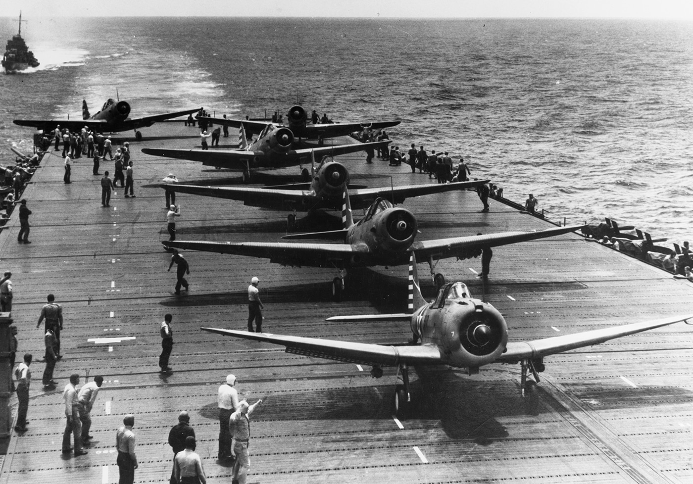 This photo shows several SBD scout bombers positioned in tight formation on the flight deck of USS Enterprise (CV-6) as they prepare for takeoff on a mission against Japanese forces. The aircraft are lined up along the deck, engines running and propellers spinning, with flight crew performing last-minute checks. Deck personnel signal to the pilots and stand ready to assist with the launch process. The sea is visible beyond the deck, underscoring the isolated setting of carrier operations. Bombs or other ordnance can be seen attached to the undersides of the aircraft, indicating the strike nature of the sortie. The image captures the intense activity and coordination required to launch multiple planes in quick succession. USS Enterprise’s air group played a pivotal role in attacking enemy ships and installations across the Pacific theater. This moment reflects the ship’s relentless operational pace during the war.