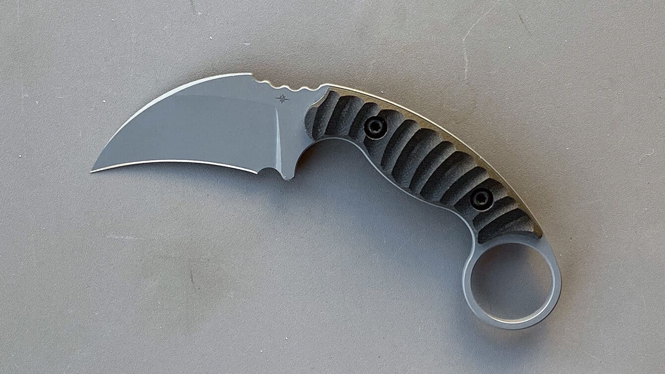 Close-up photo of the Toor Knives Karsumba R’s 2½-inch hawkbill blade made from Crucible CPM M4 tool steel. The curved profile and full flat grind are visible, emphasizing the knife’s cutting geometry. The matte KG GunKote finish gives the blade a subdued tactical look while protecting it from wear and corrosion. Fine details like the spine jimping and aggressive tip reflect Toor’s focus on precision and functionality. The image highlights the balance between compact size and heavy-duty performance that defines the Karsumba R. It visually supports the review’s section on materials and edge quality.